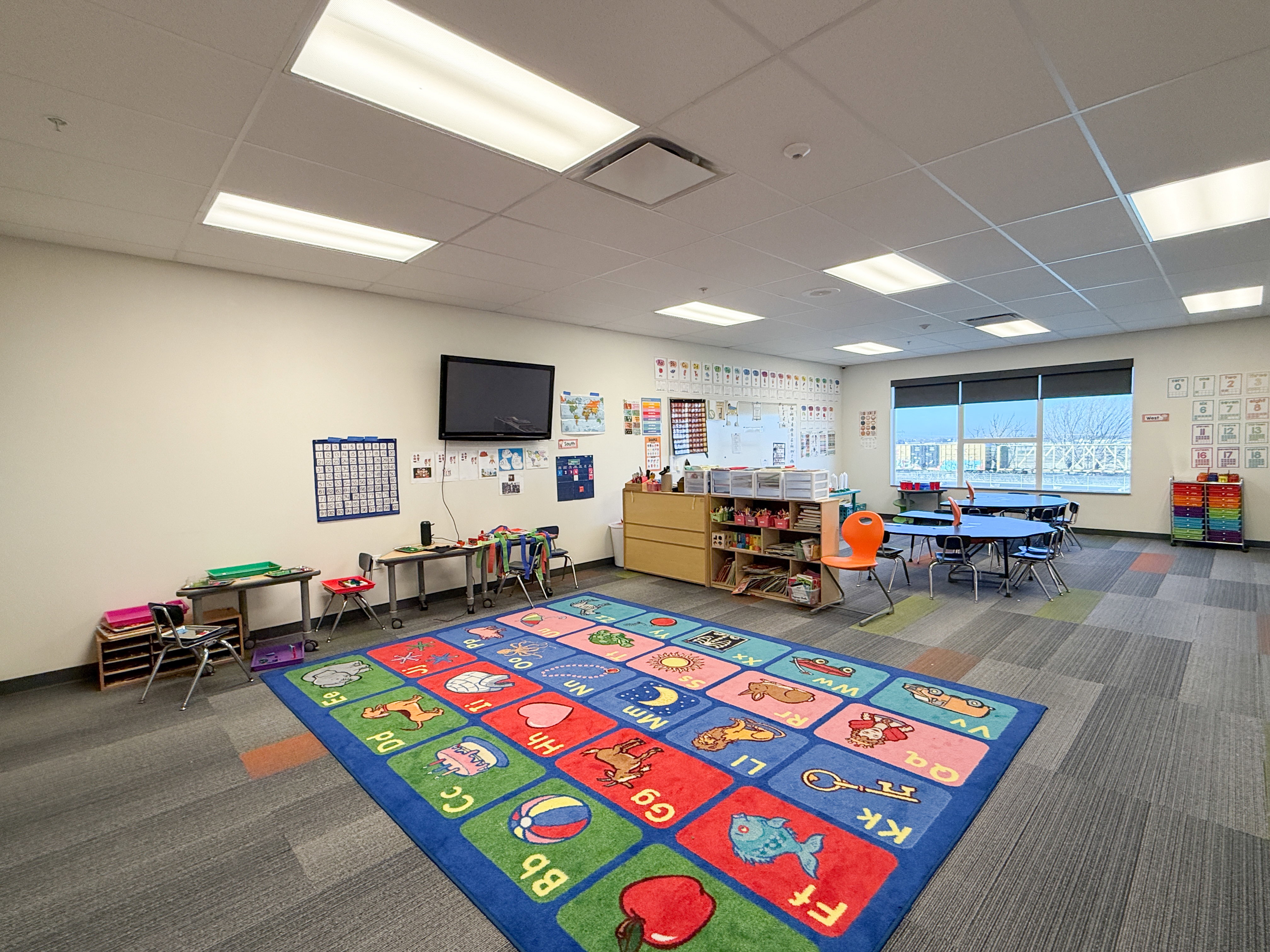 Spacious room featuring modular carpet flooring, ample overhead lighting, and a large window with roller shades