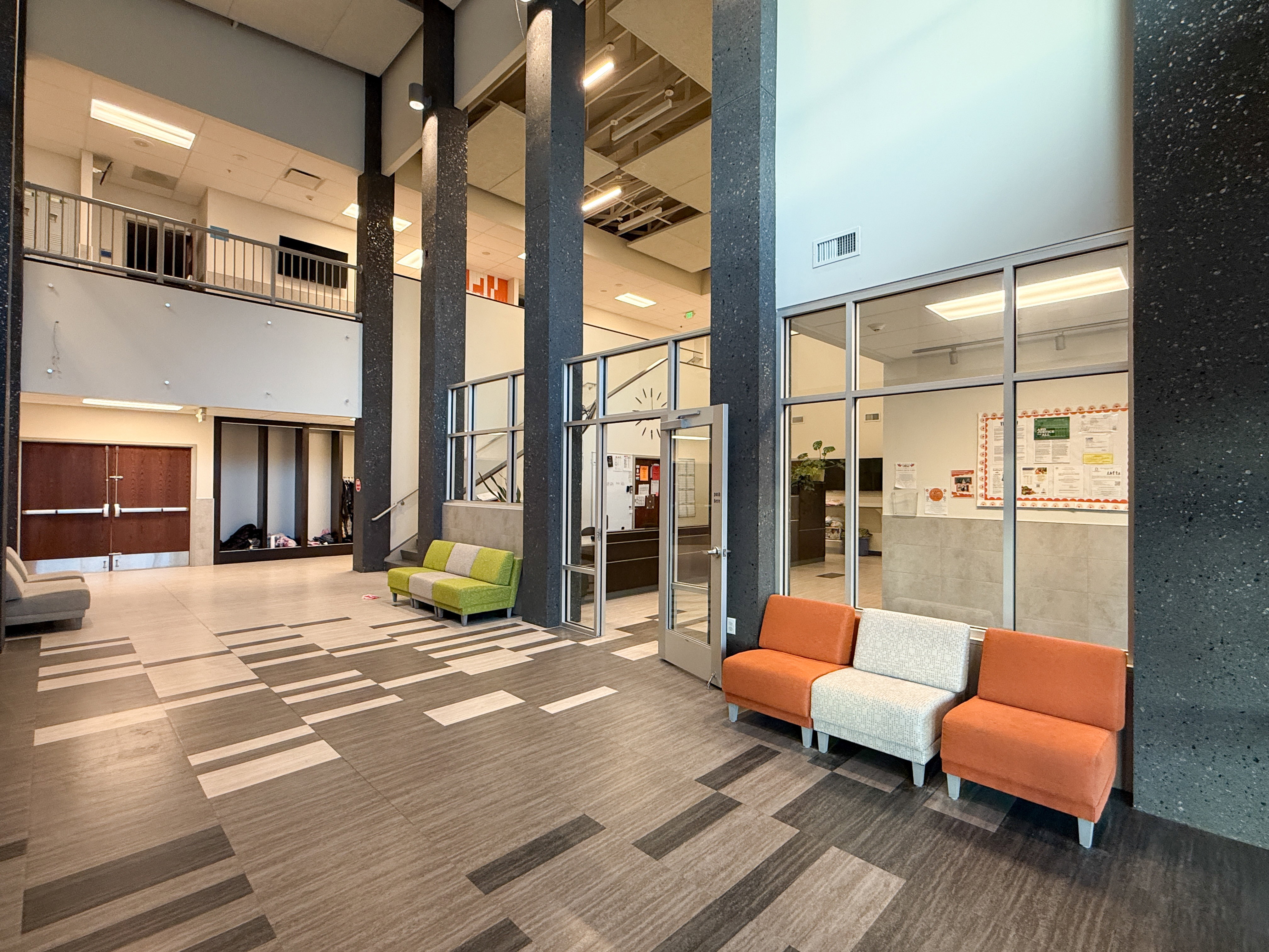 Two-story lobby featuring a patterned tile floor, prominent dark textured pillars, a glass-paneled office area, and double wood-finish doors