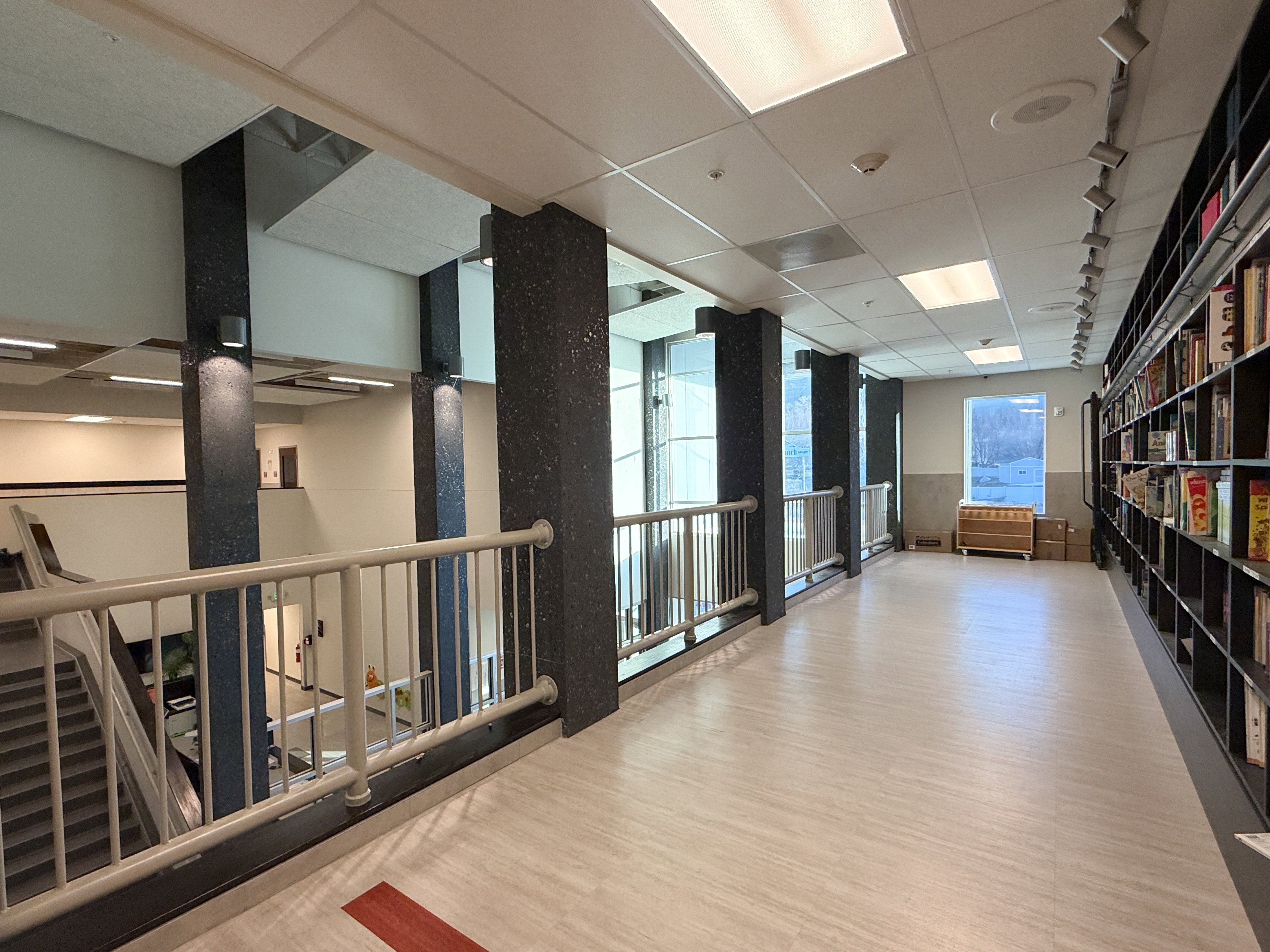 Upper-level hallway featuring light wood-finish flooring, a full-height built-in shelving unit, and a metal railing overlooking a lower level
