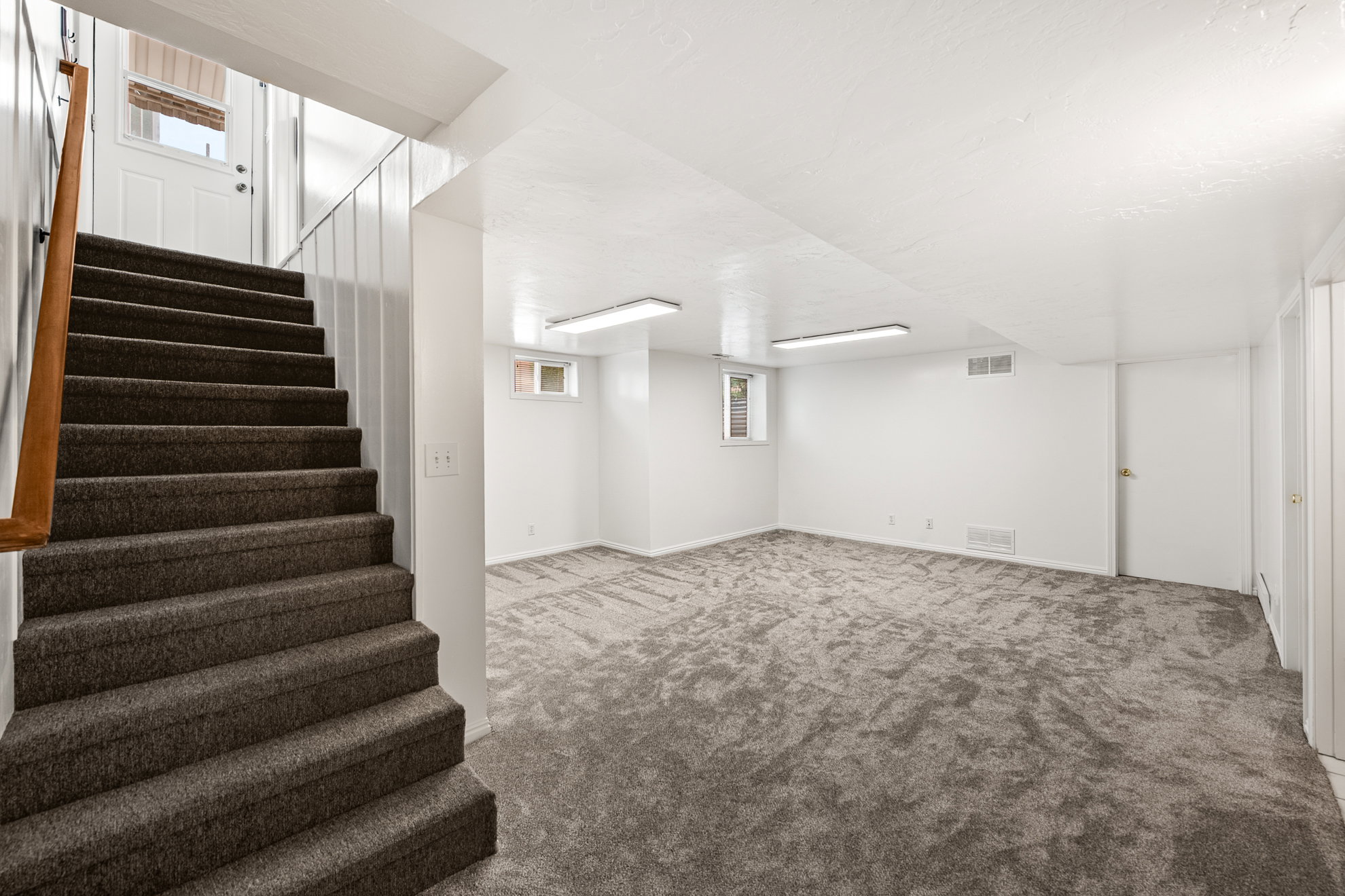 Spacious lower-level room featuring plush gray carpeting, white painted walls, and overhead fluorescent lighting