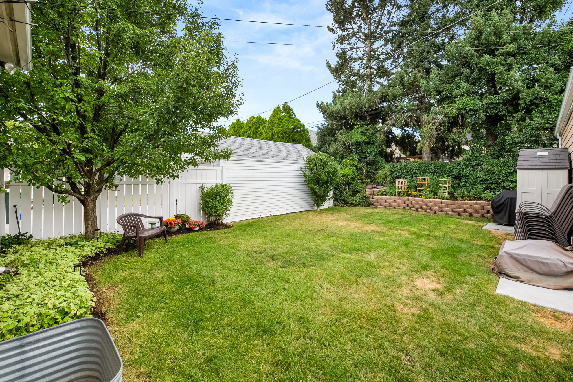 Private backyard featuring a manicured lawn, white picket fencing, mature trees, and a tiered retaining wall