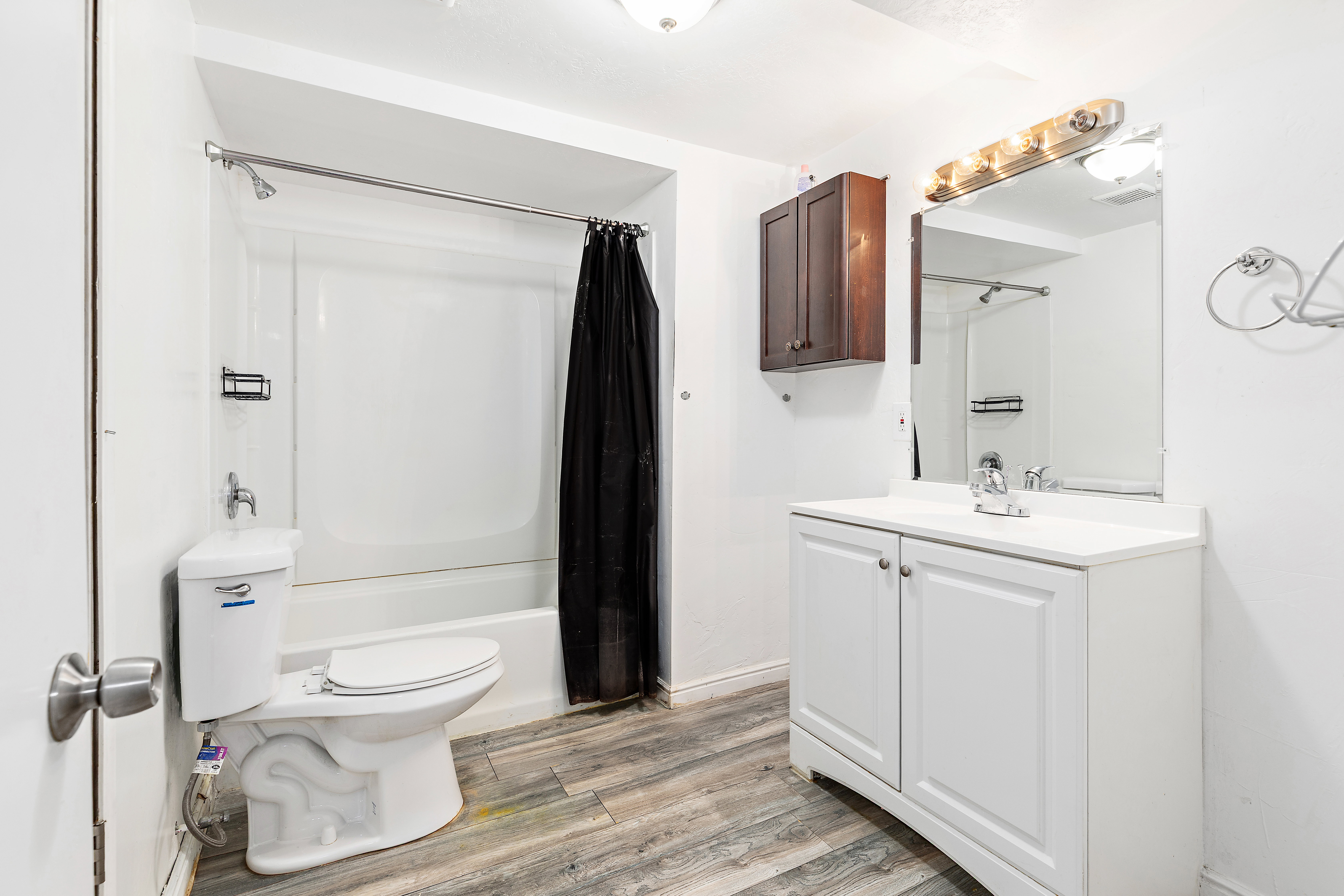 Bathroom featuring a white vanity with integrated sink, a rectangular mirror with a multi-bulb light fixture, a shower-tub combination with a white surround, and wood-finish flooring
