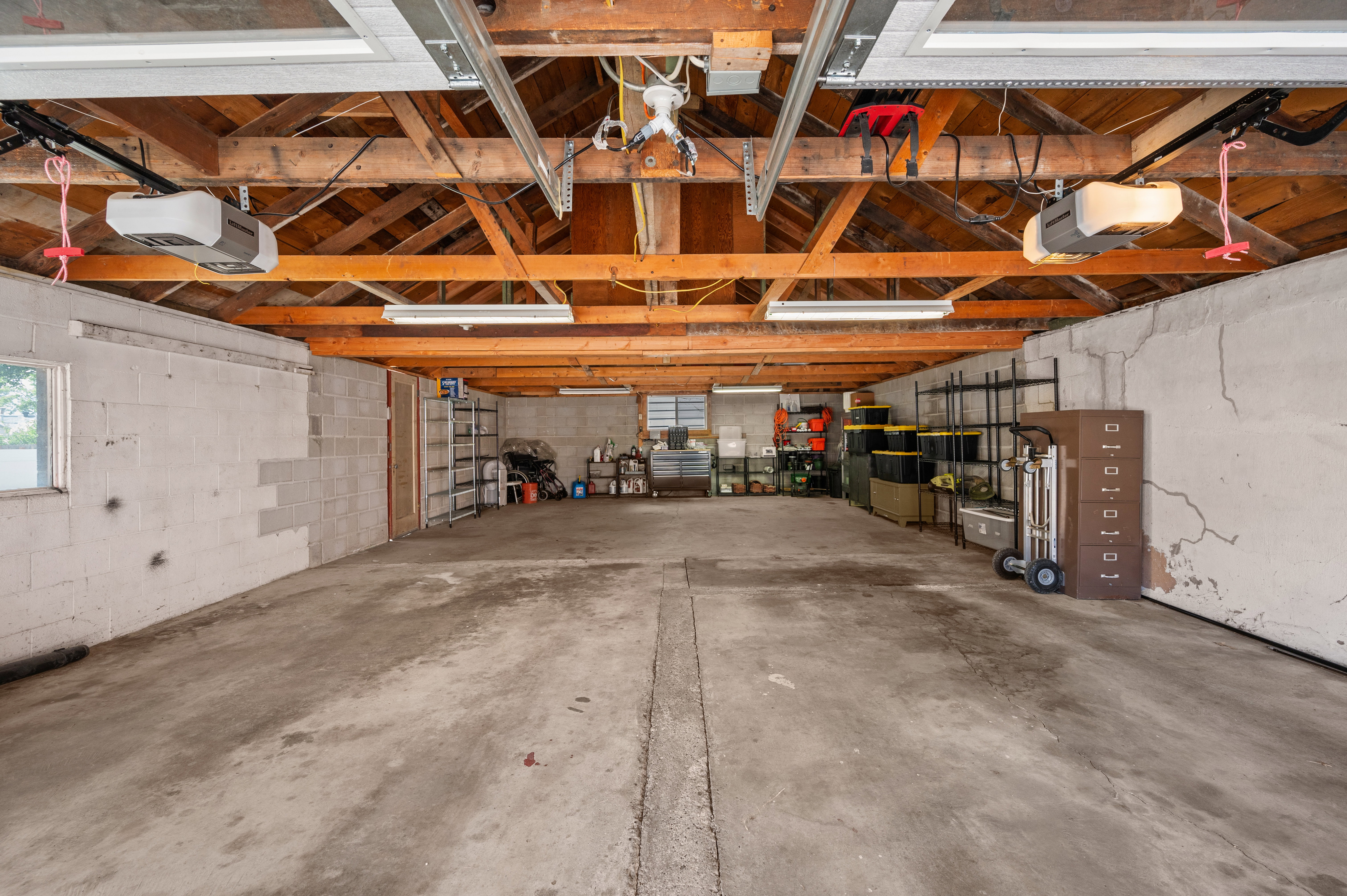 Spacious garage featuring concrete flooring, exposed wood beam ceiling, two automatic garage door openers, and fluorescent lighting fixtures