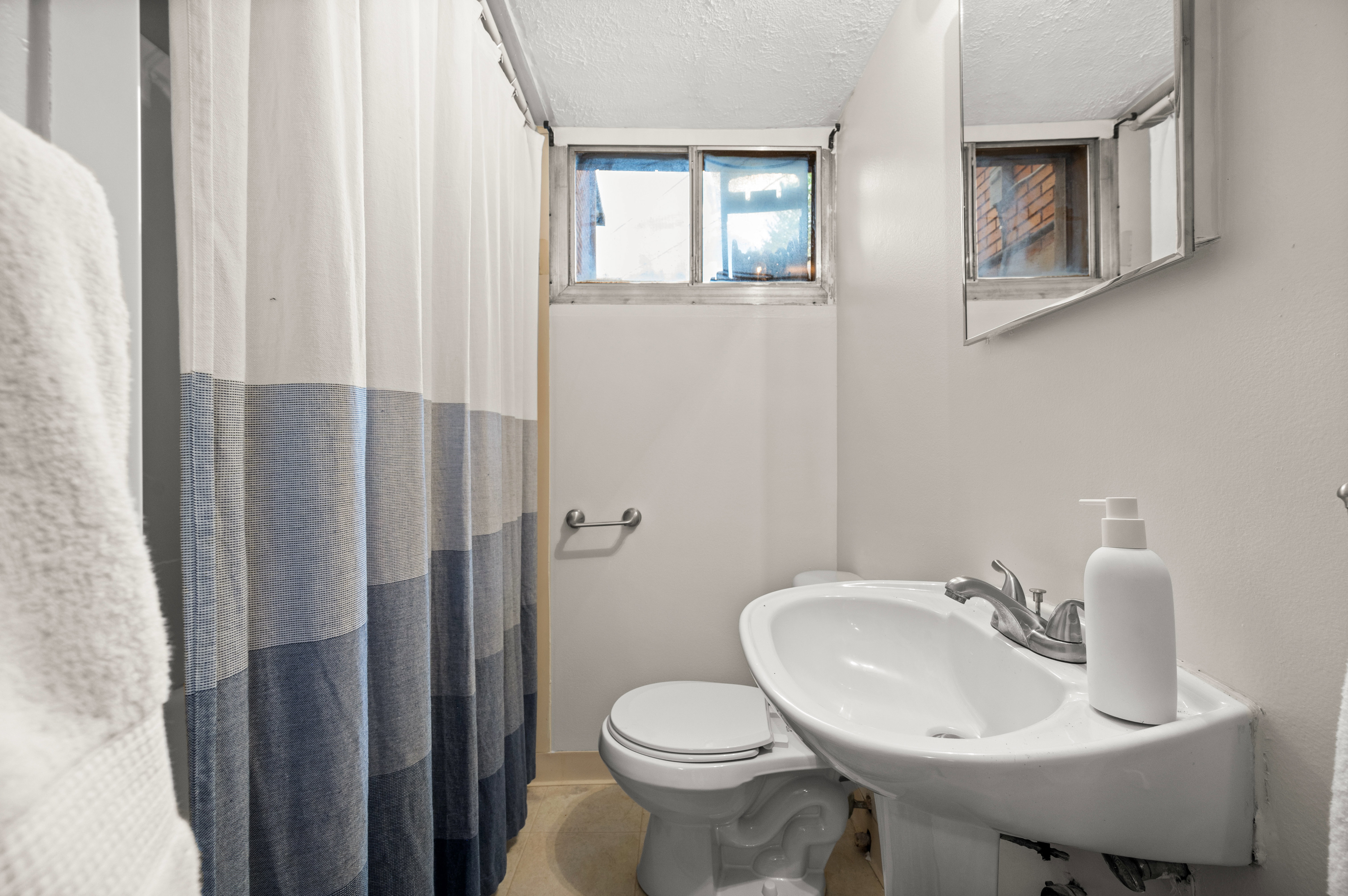 Compact bathroom featuring a wall-mounted pedestal sink with a chrome faucet, a toilet, a small window, and a wall-mounted medicine cabinet with a mirror