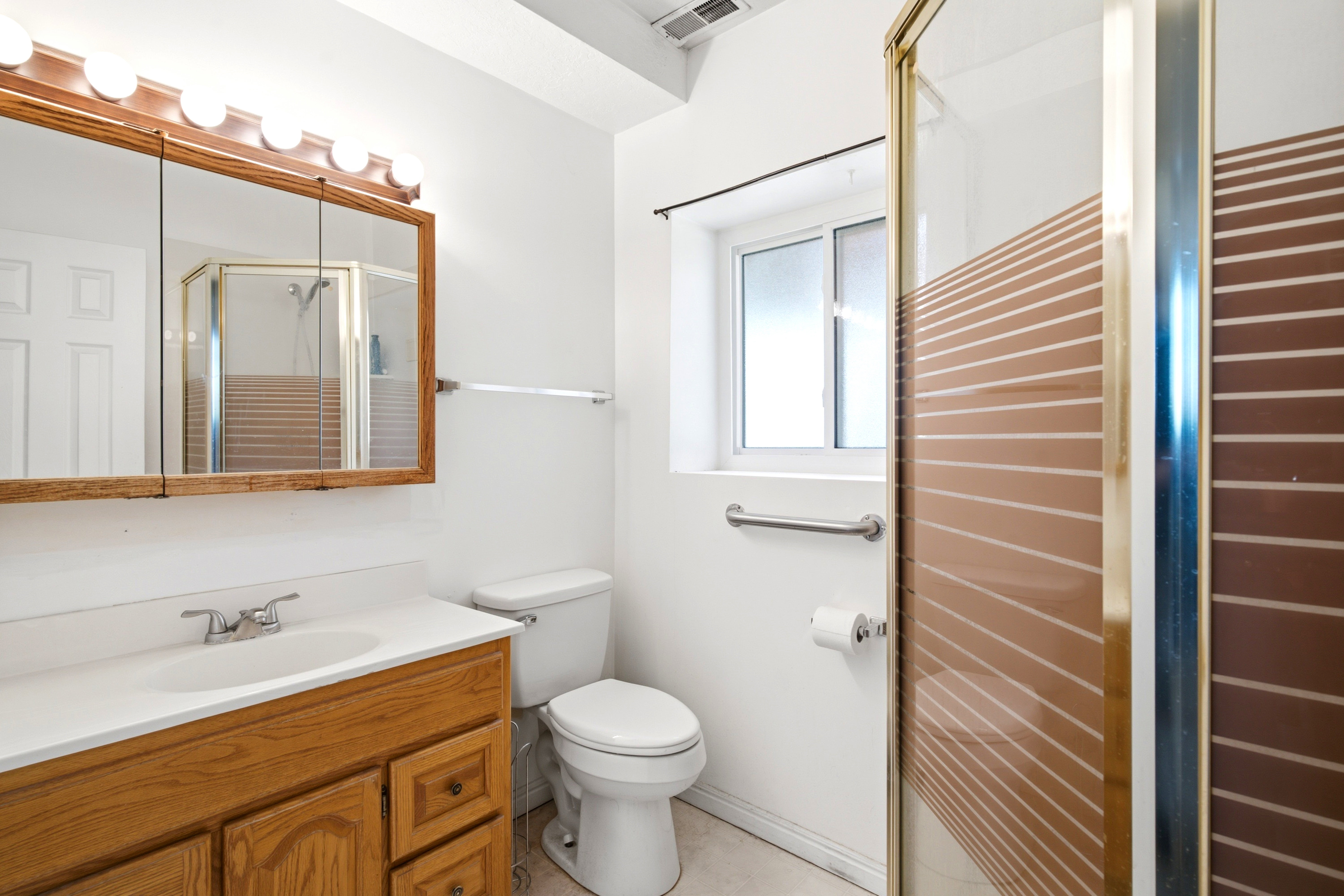 Bathroom featuring a wood-finish vanity with a white countertop and integrated sink, a mirrored medicine cabinet with overhead globe lighting, and a shower enclosure with a patterned glass panel and a gold-tone frame
