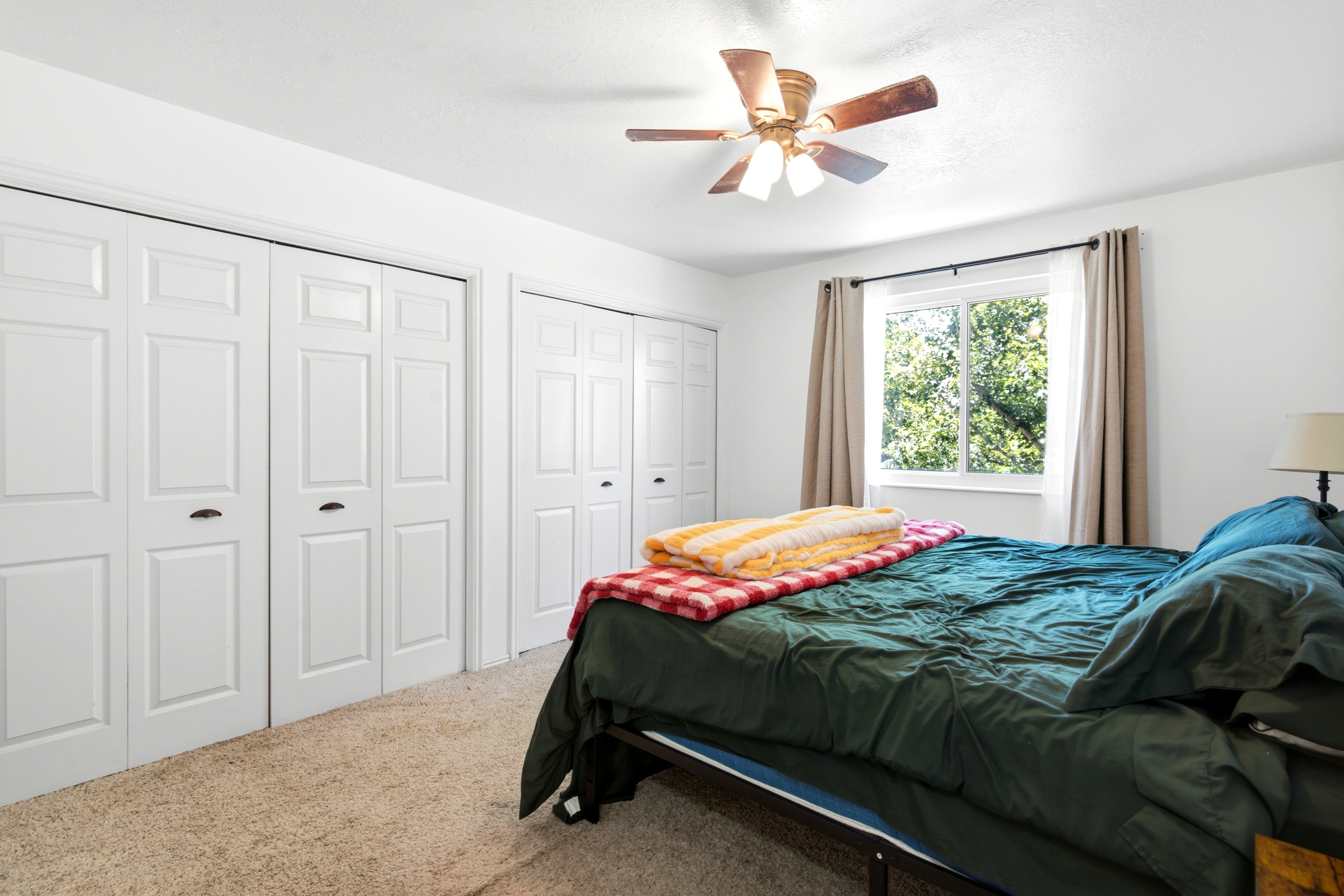White paneled bi-fold closet doors, neutral tone carpeting, ceiling fan with integrated lighting, and a window with exterior tree scenery