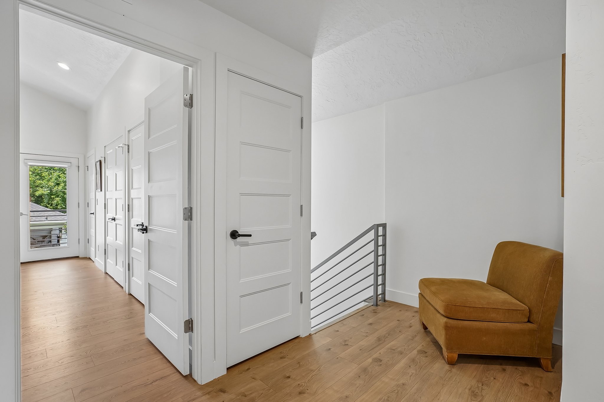 Hallway featuring light wood-finish flooring, white paneled doors with black hardware, recessed lighting, and a contemporary metal railing