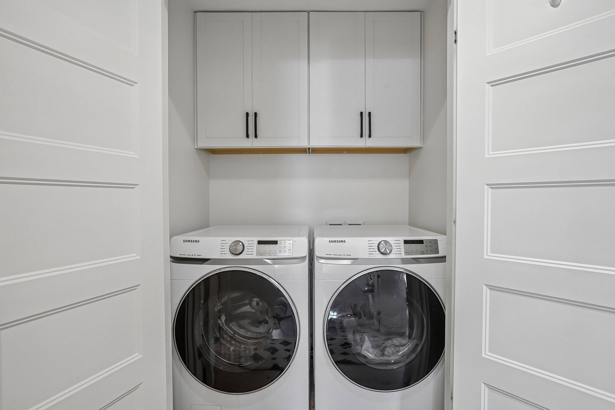 Dedicated laundry closet featuring white paneled doors, white upper cabinetry with black hardware, and a wood shelf