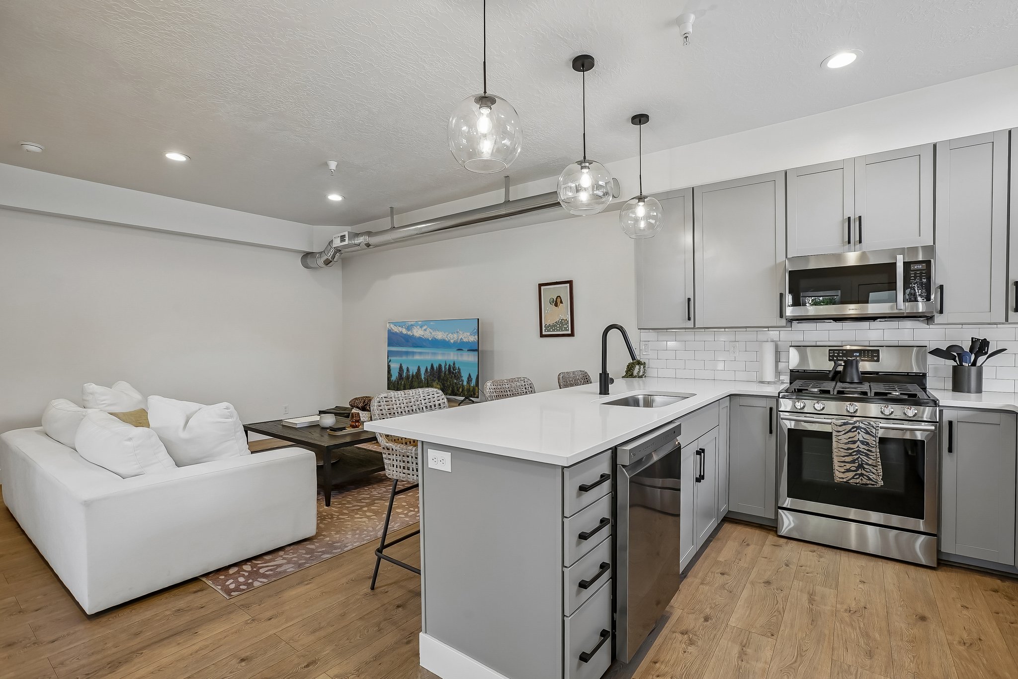 Open-concept kitchen featuring an island with white countertop, grey cabinetry with black hardware, stainless steel appliances, and wood-finish flooring