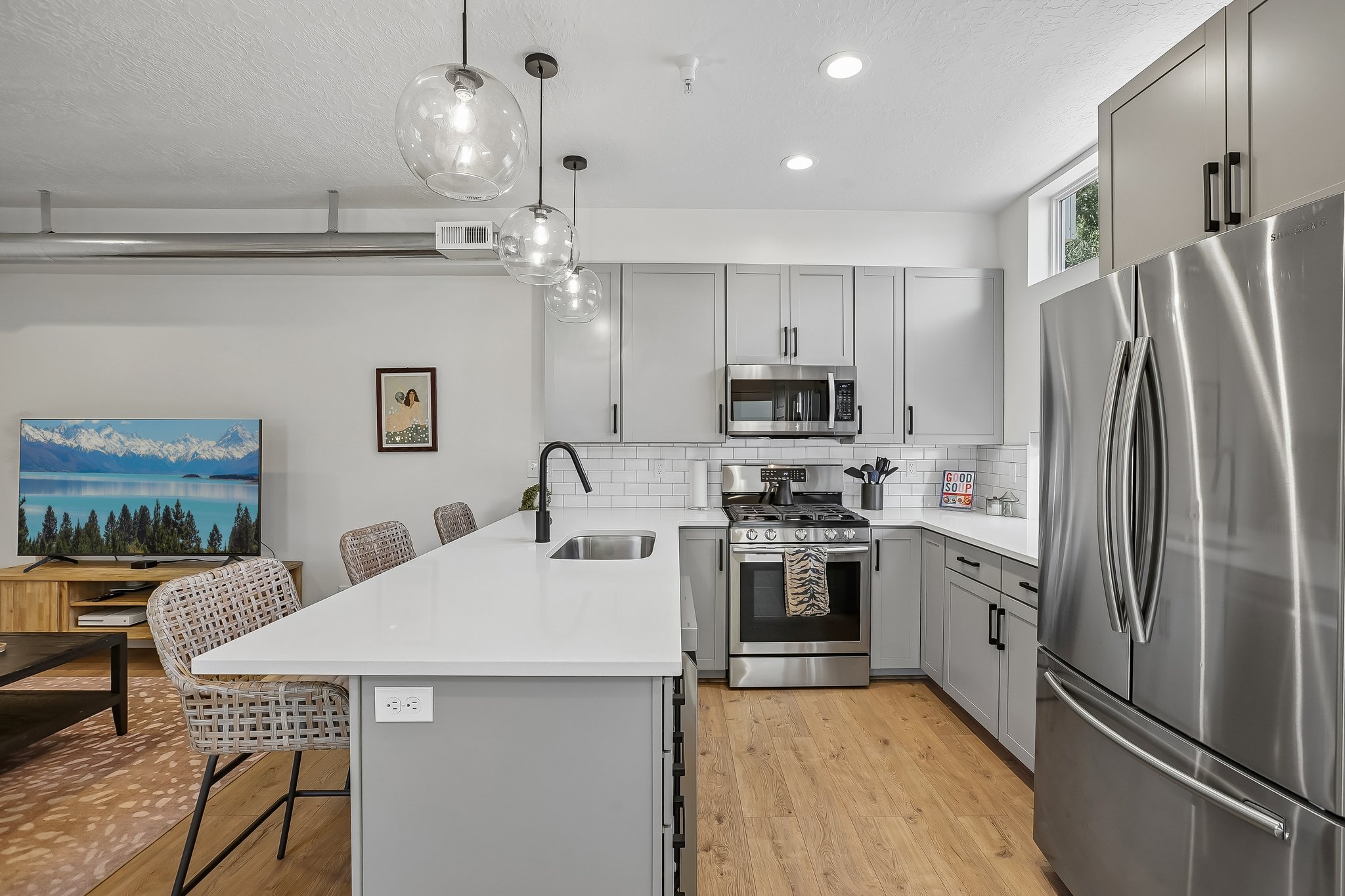 Modern kitchen featuring light gray cabinetry, stainless steel appliances, white subway tile backsplash, a large kitchen island with a white countertop and undermount sink, and wood-finish flooring