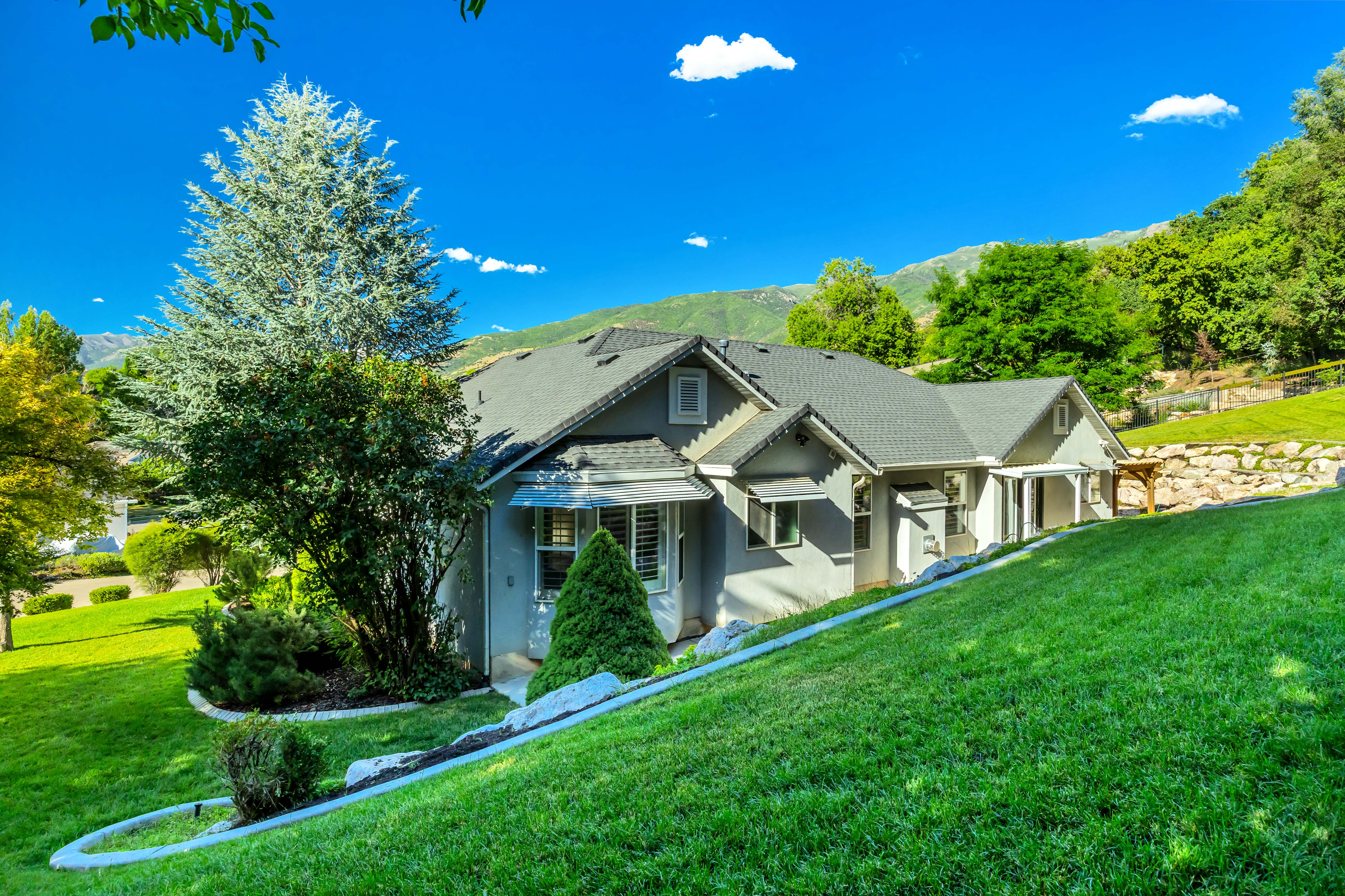 Sloped lot featuring a gray exterior residence with a dark shingle roof, white trim details, and multiple windows with louvered shutters
