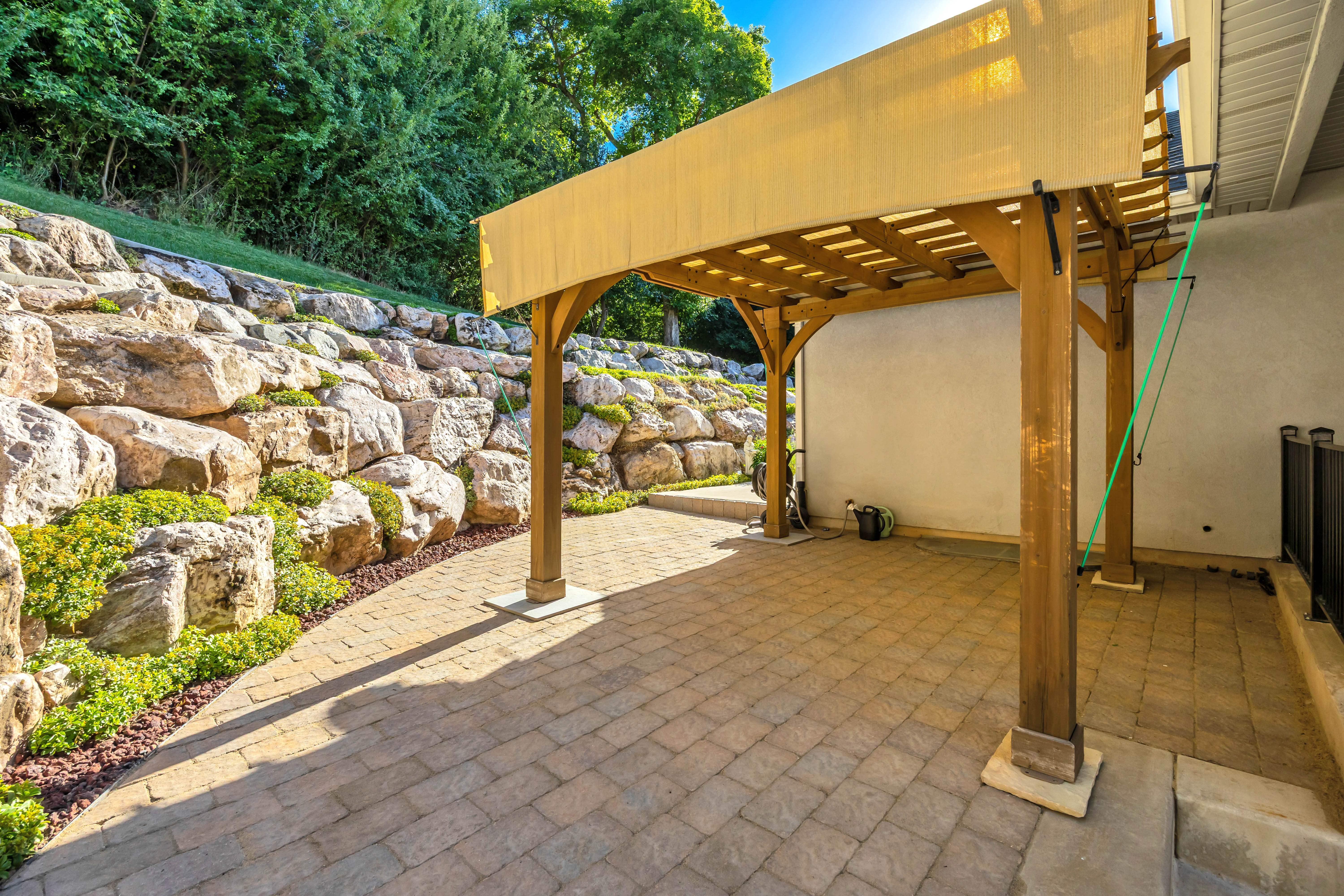 Paver patio featuring a wood pergola with a sunshade, set against a tiered rock retaining wall with integrated landscaping