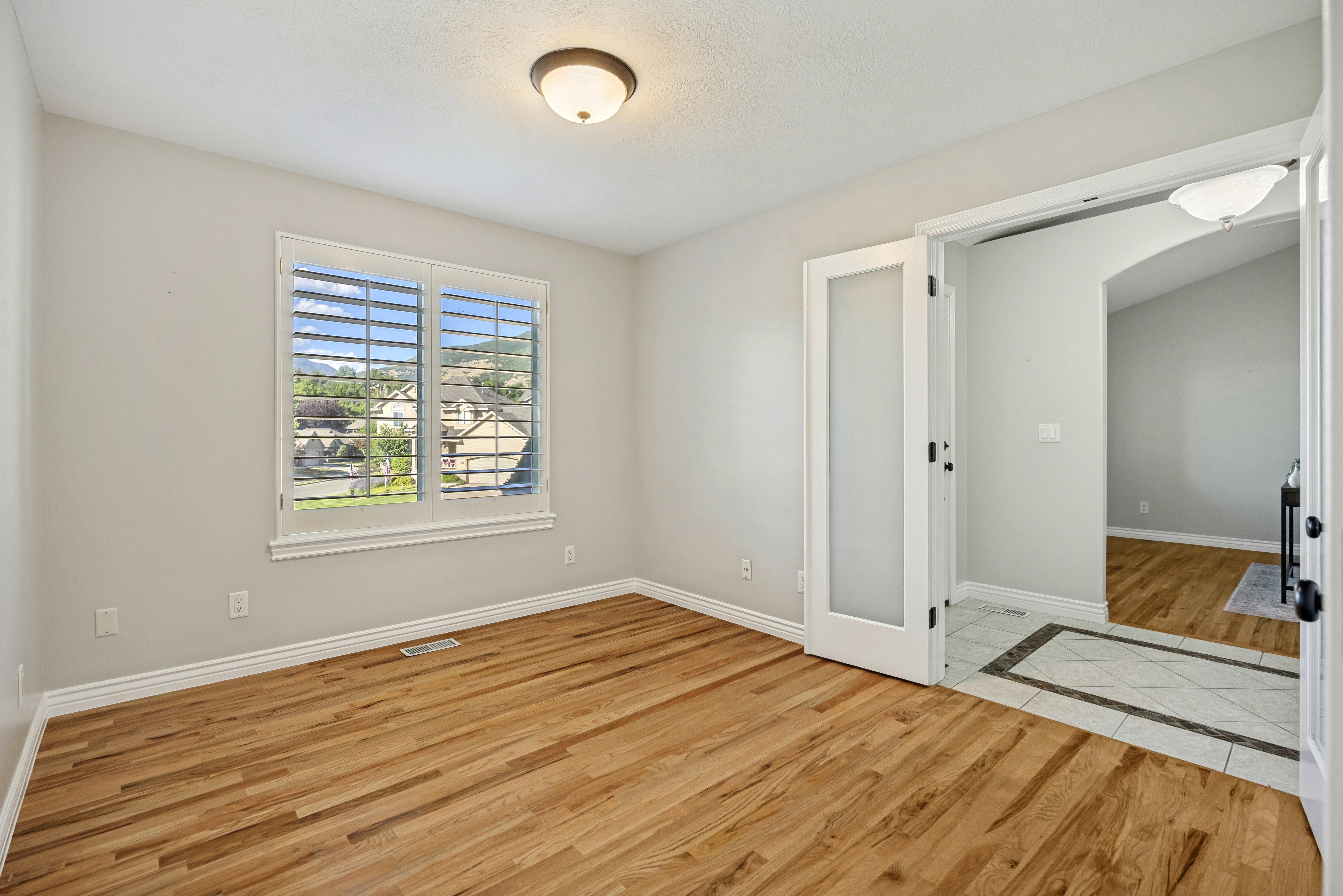 2nd Bedroom or flex room that can be an office if desired. Entails a large window with plantation shutters, a contemporary ceiling light fixture, and double French doors with frosted glass panels leading to a tiled entryway