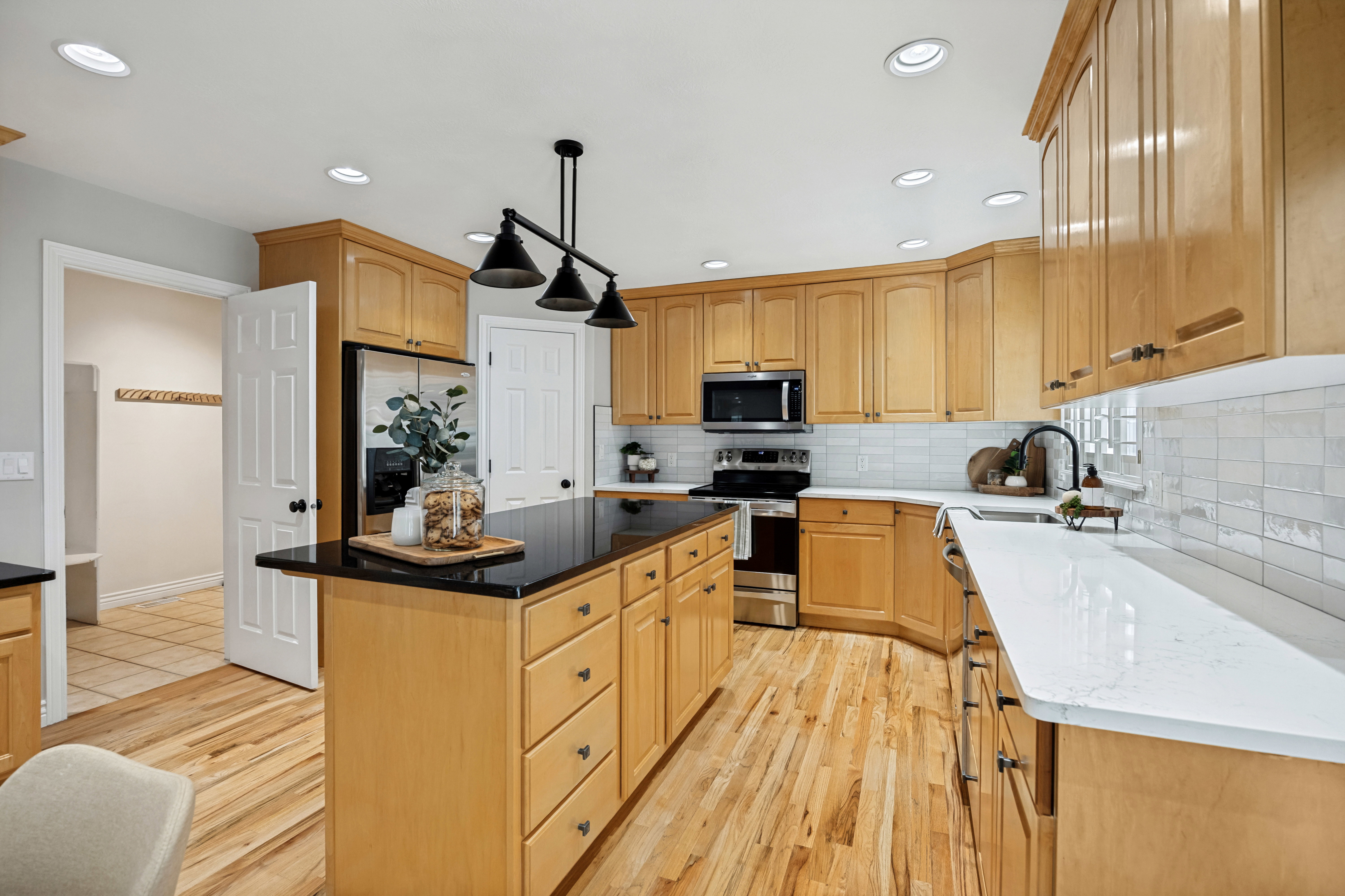 Kitchen featuring wood flooring, light custom wood cabinetry, a central island with a dark quartz countertop, white perimeter quartz countertops, and a white tile backsplash