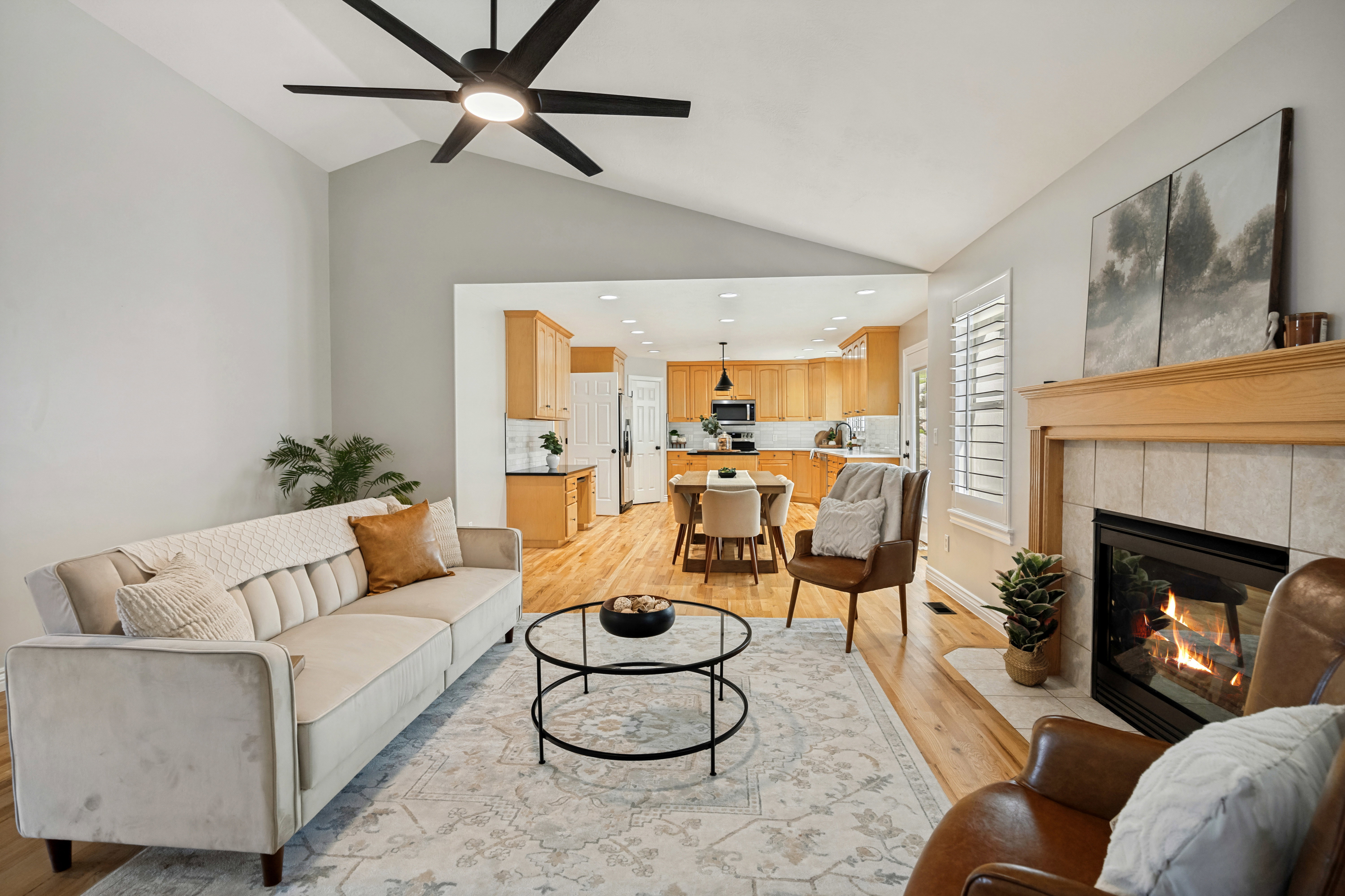 Vaulted ceilings with a ceiling fan, hardwood flooring, fireplace with a tiled surround and wood mantel, plantation shutters, and recessed lighting in the kitchen