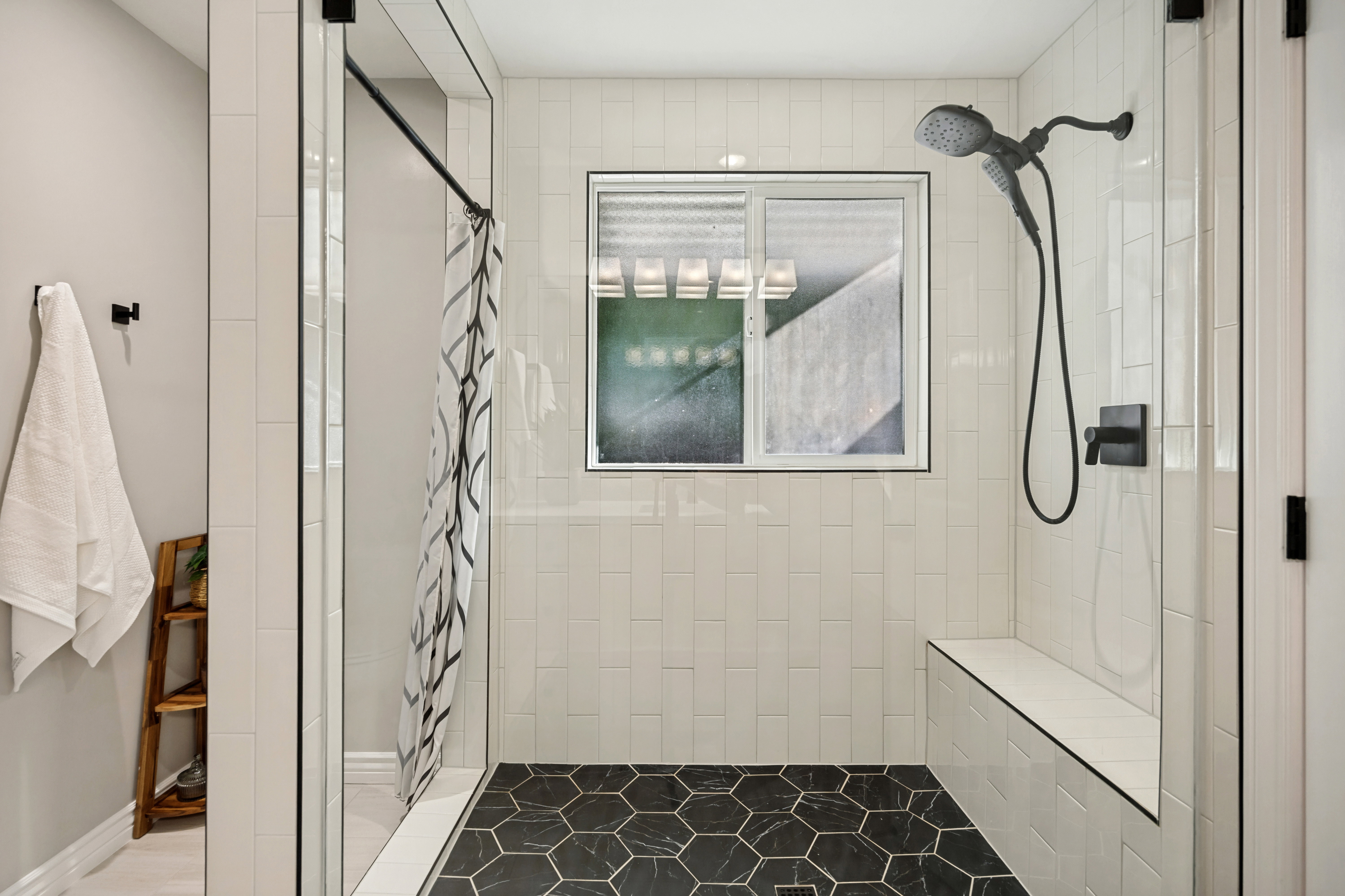 LARGE and BRIGHT Walk-in shower featuring black hexagonal floor tiles, vertical white subway wall tiles, a built-in bench, black fixtures, and frameless glass