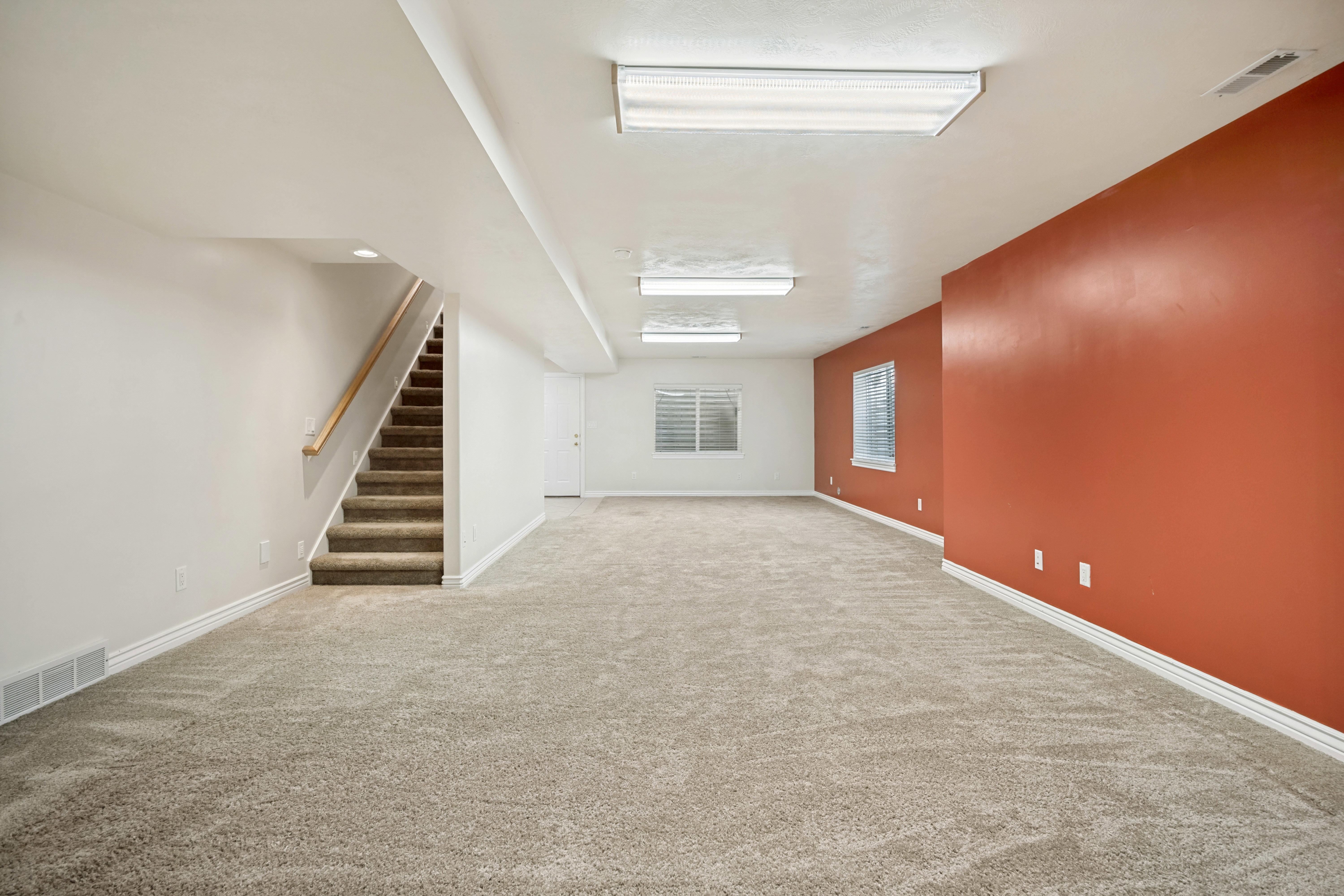 Spacious lower level family room featuring neutral carpeting, white baseboards, and a contrasting accent wall. Opens to the kitchenette and can have dining, living, and game room space.