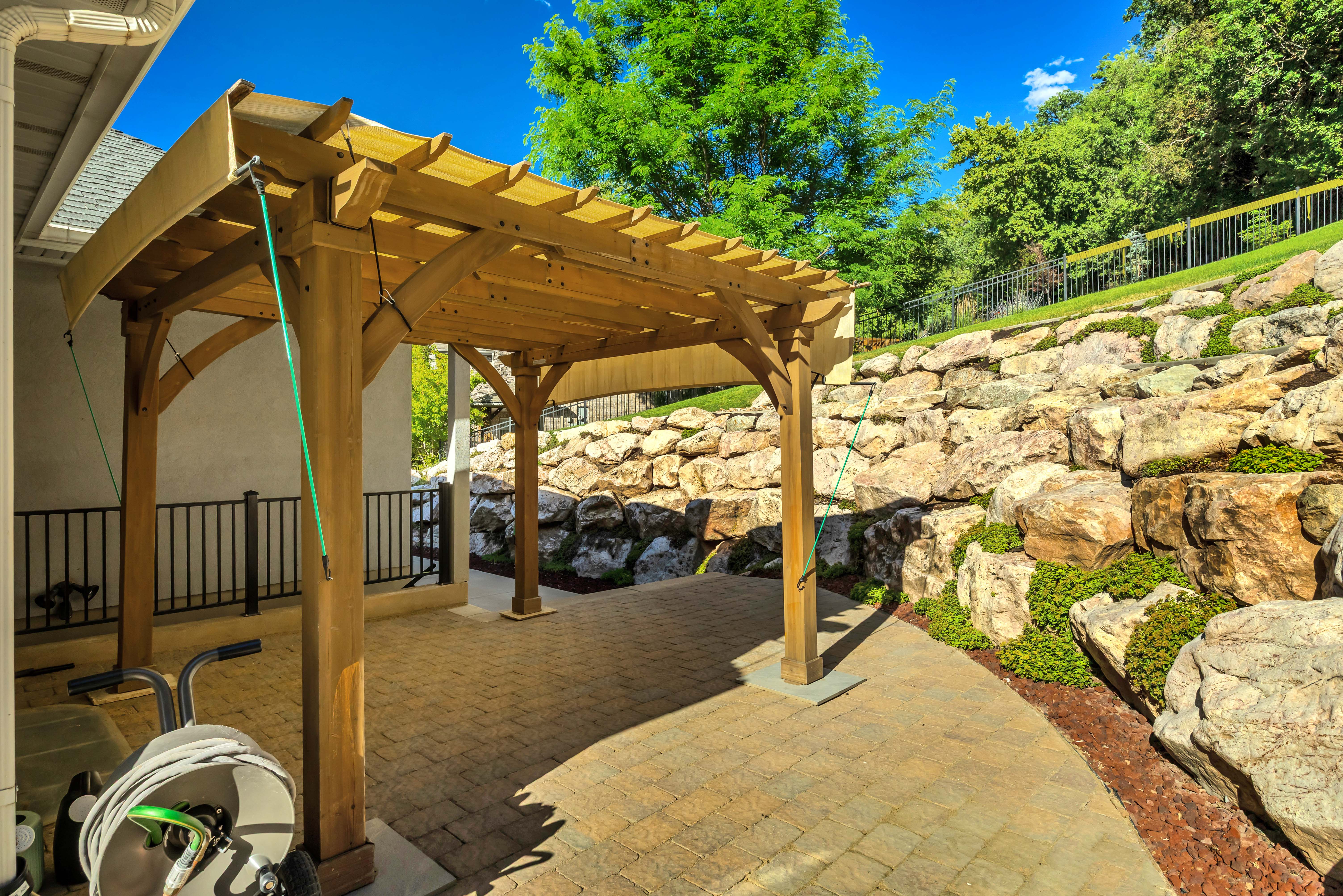 Expansive patio featuring a wooden pergola with a fabric canopy, paver flooring, and a stone retaining wall with integrated plantings
