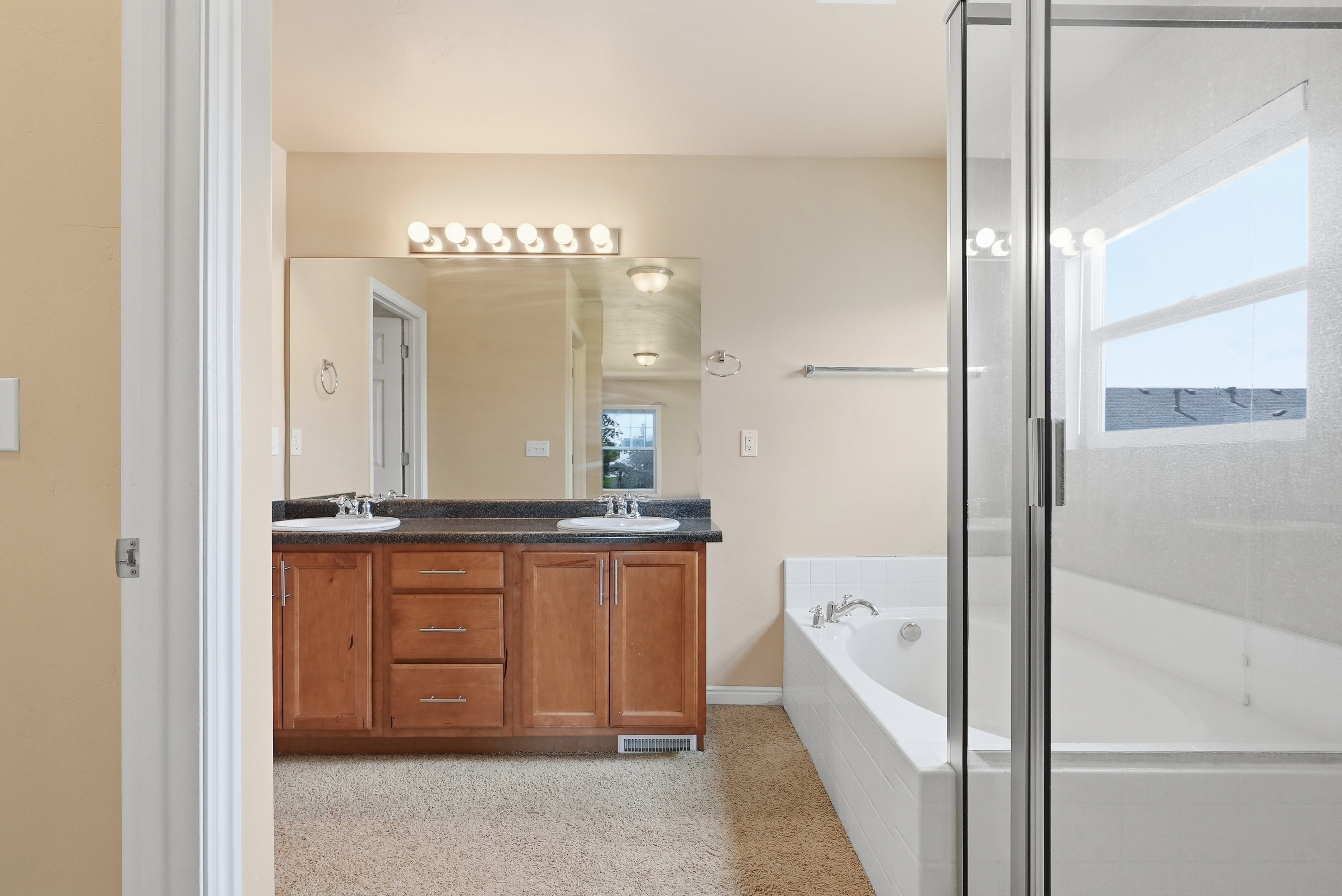 Bathroom featuring a double vanity with wood cabinetry and a dark countertop, an expansive mirror with strip lighting, a built-in soaking tub, and a separate glass-enclosed shower