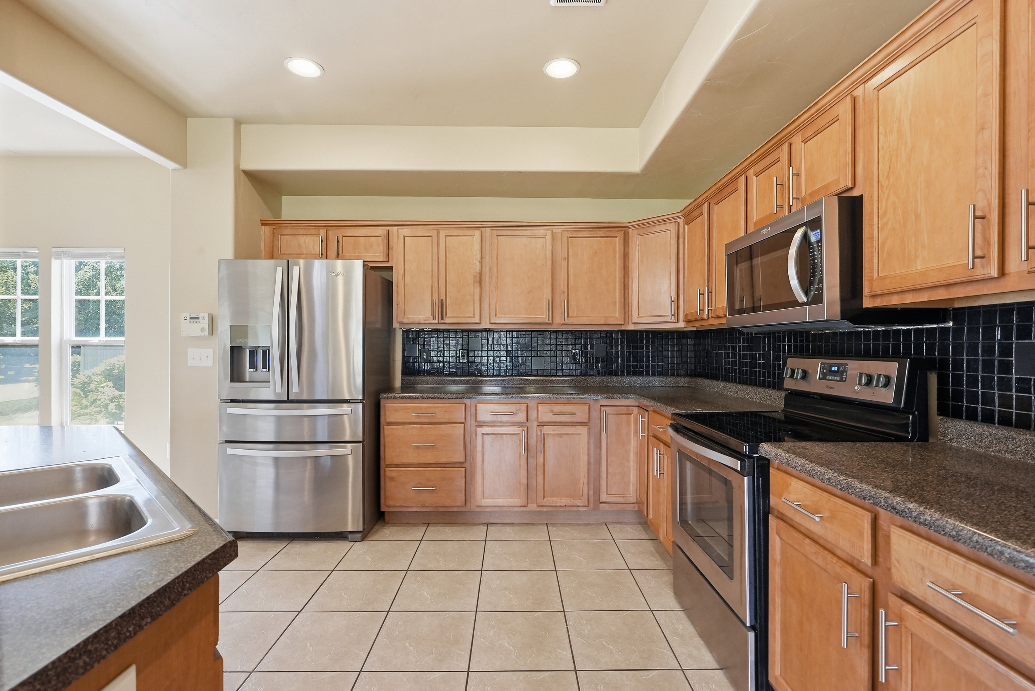 Kitchen featuring light wood cabinetry, stainless steel appliances, dark textured countertops, black tile backsplash, and light-colored tile flooring