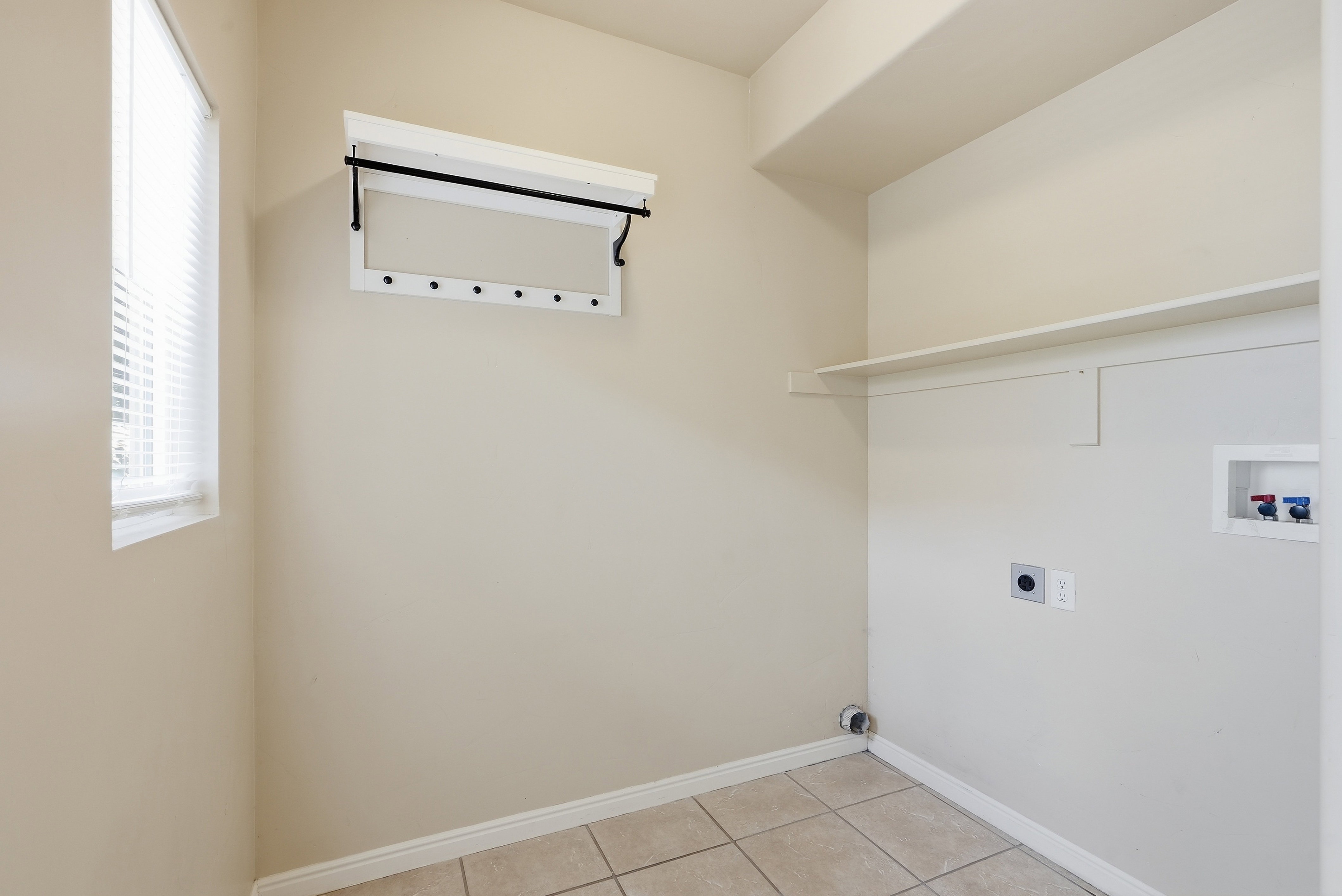 Dedicated laundry area featuring a window with blinds, a wall-mounted drying rack, a utility shelf, and tile flooring