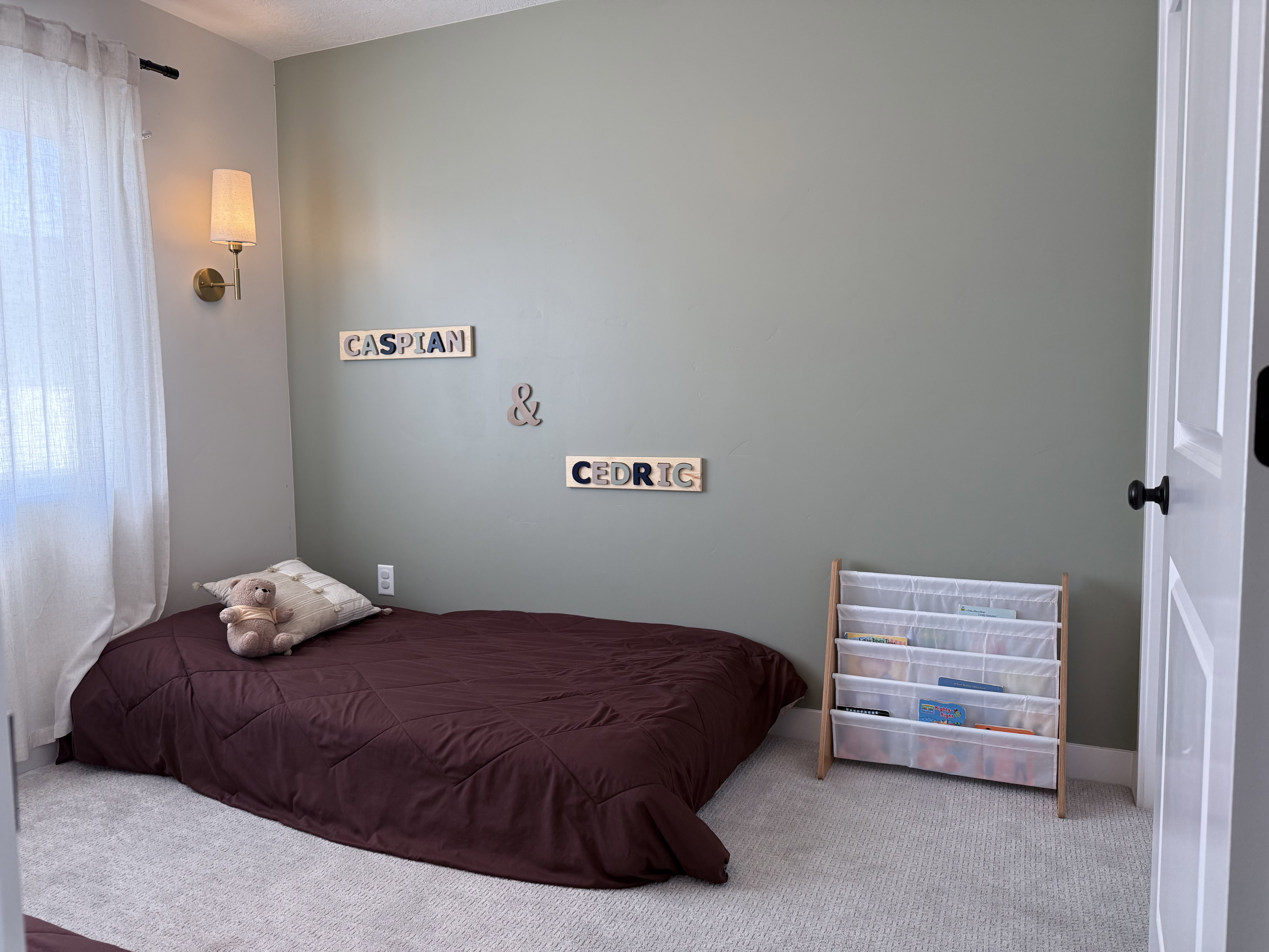Carpeted room featuring a window with sheer white curtains, a wall-mounted sconce with a fabric shade and brass-tone base, and a white paneled door with a black knob