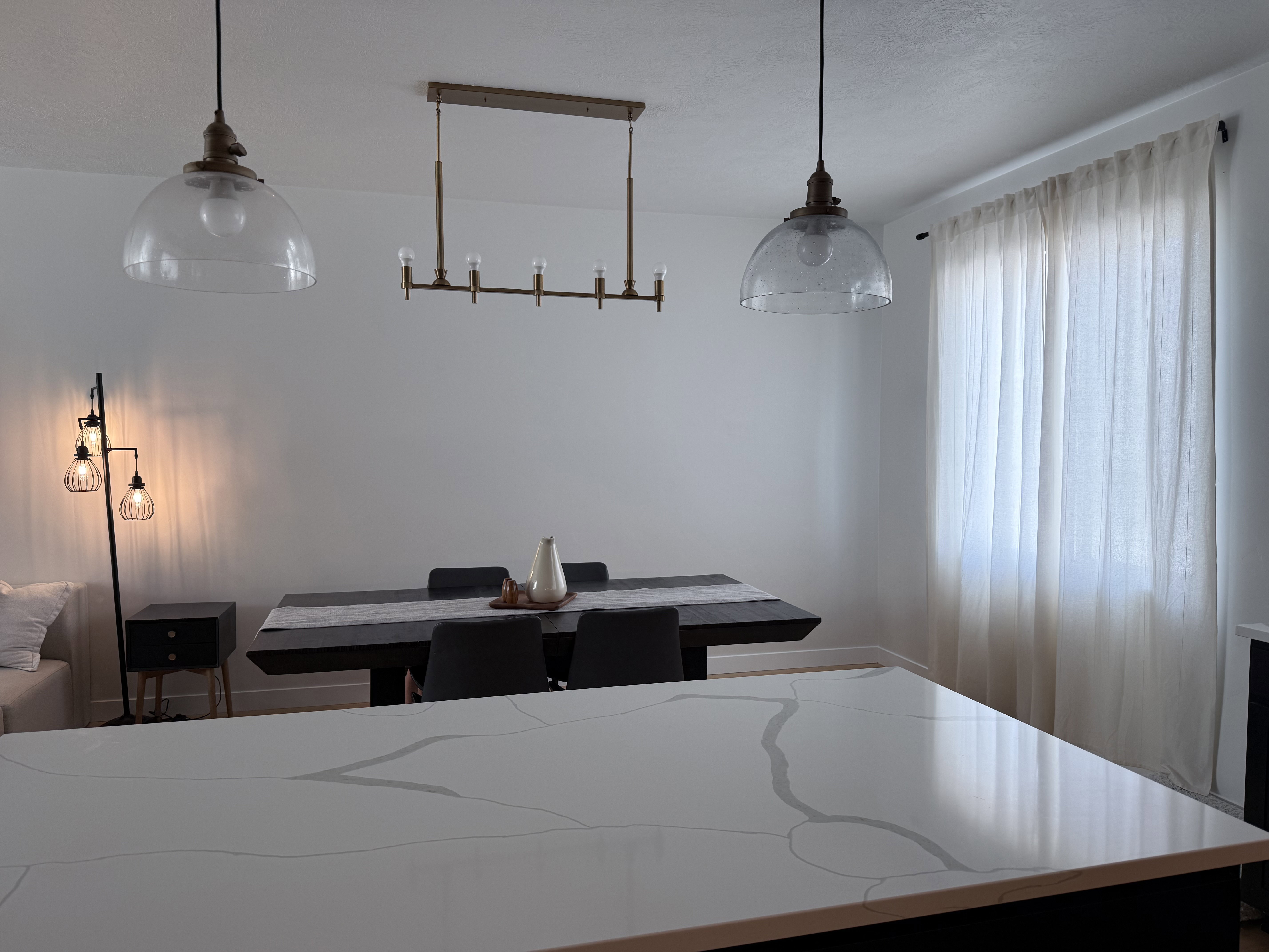 Spacious room featuring a large dining table with seating, two clear glass pendant lights, a linear chandelier, and a prominent window with sheer curtains