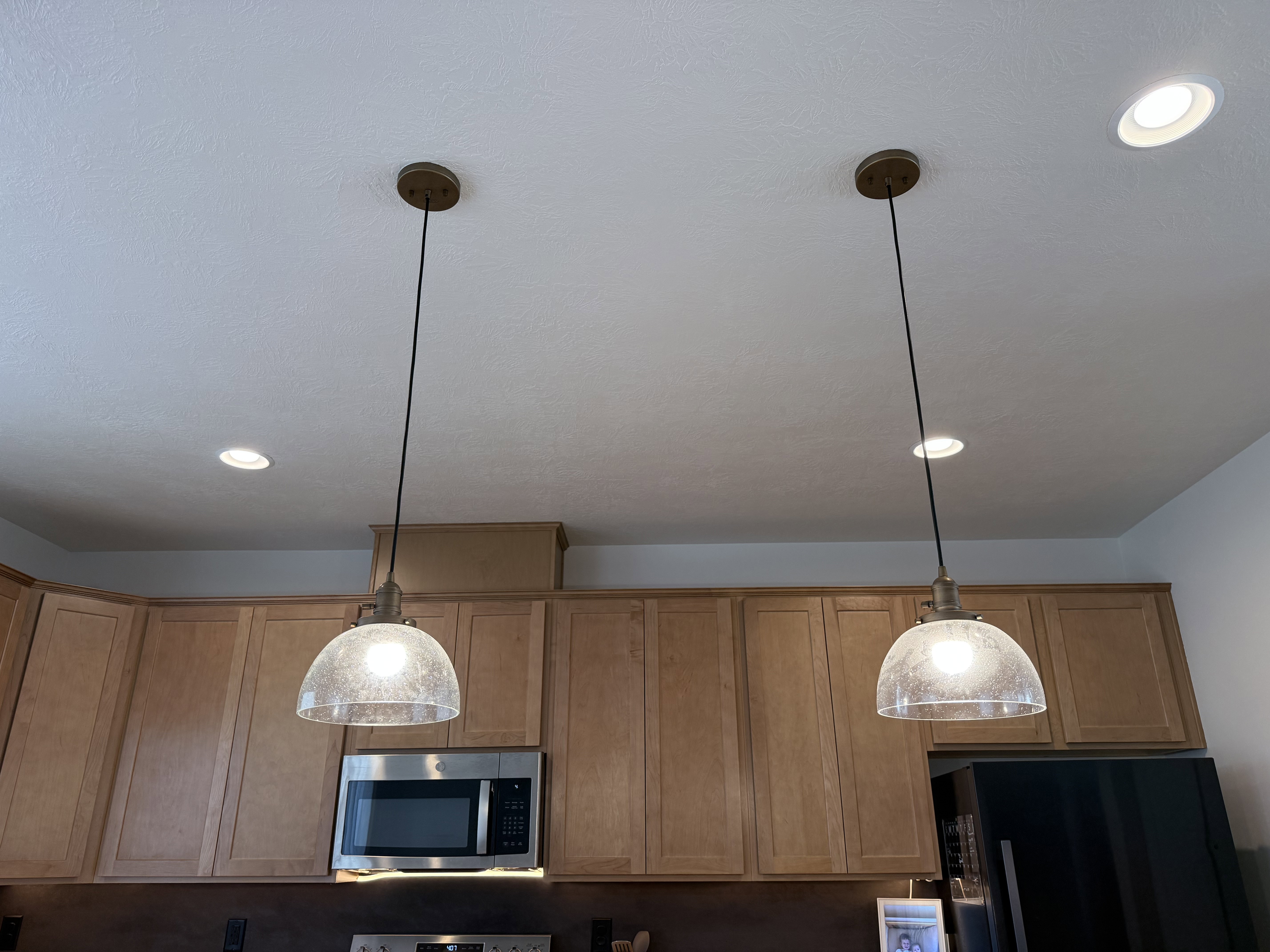 Kitchen featuring two suspended pendant lights with seeded glass shades, recessed ceiling lighting, light wood-finish cabinetry, and stainless steel appliances including a microwave