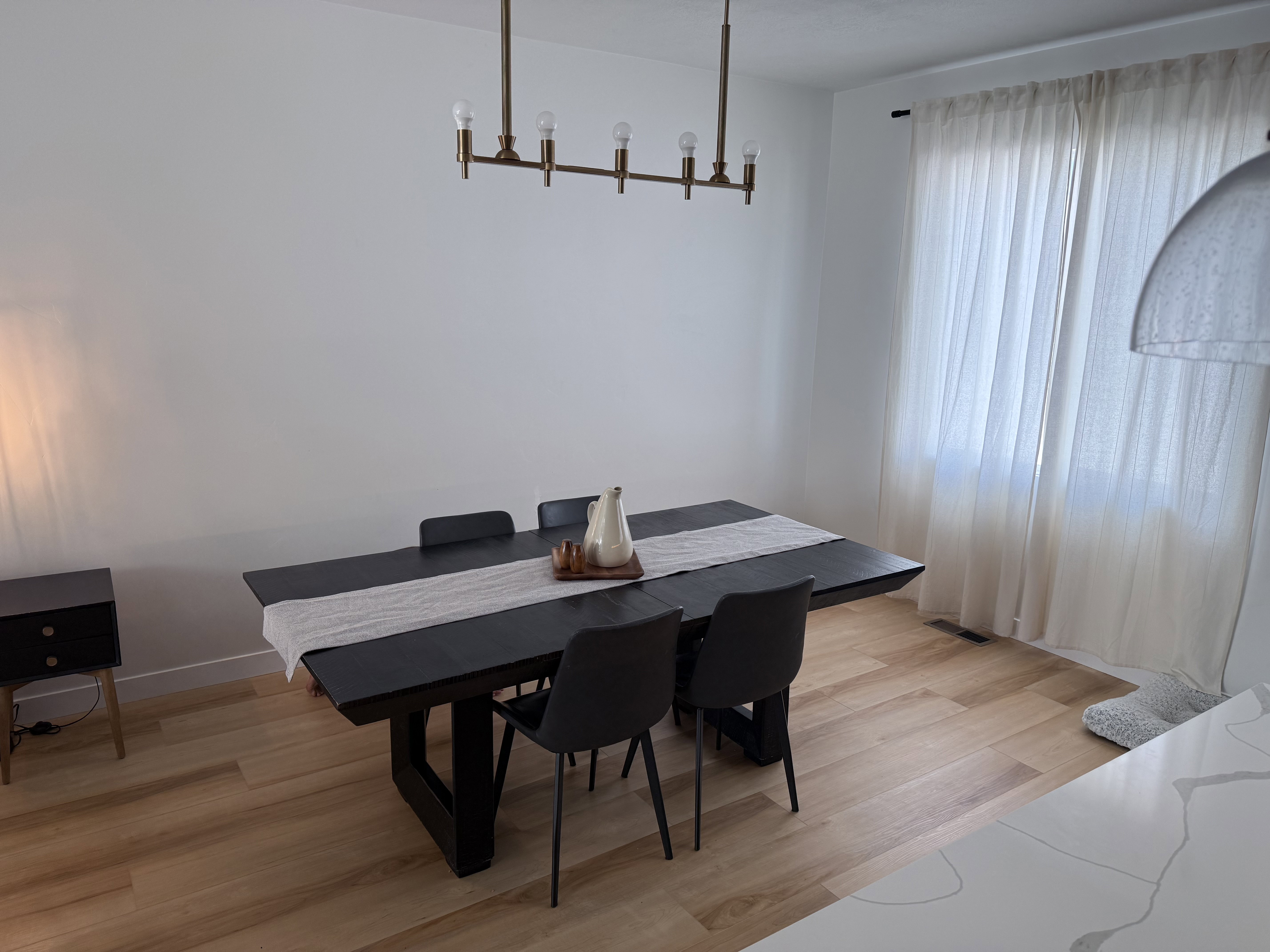 Dining area featuring light wood-finish flooring, a modern linear chandelier, and a large window with privacy curtains