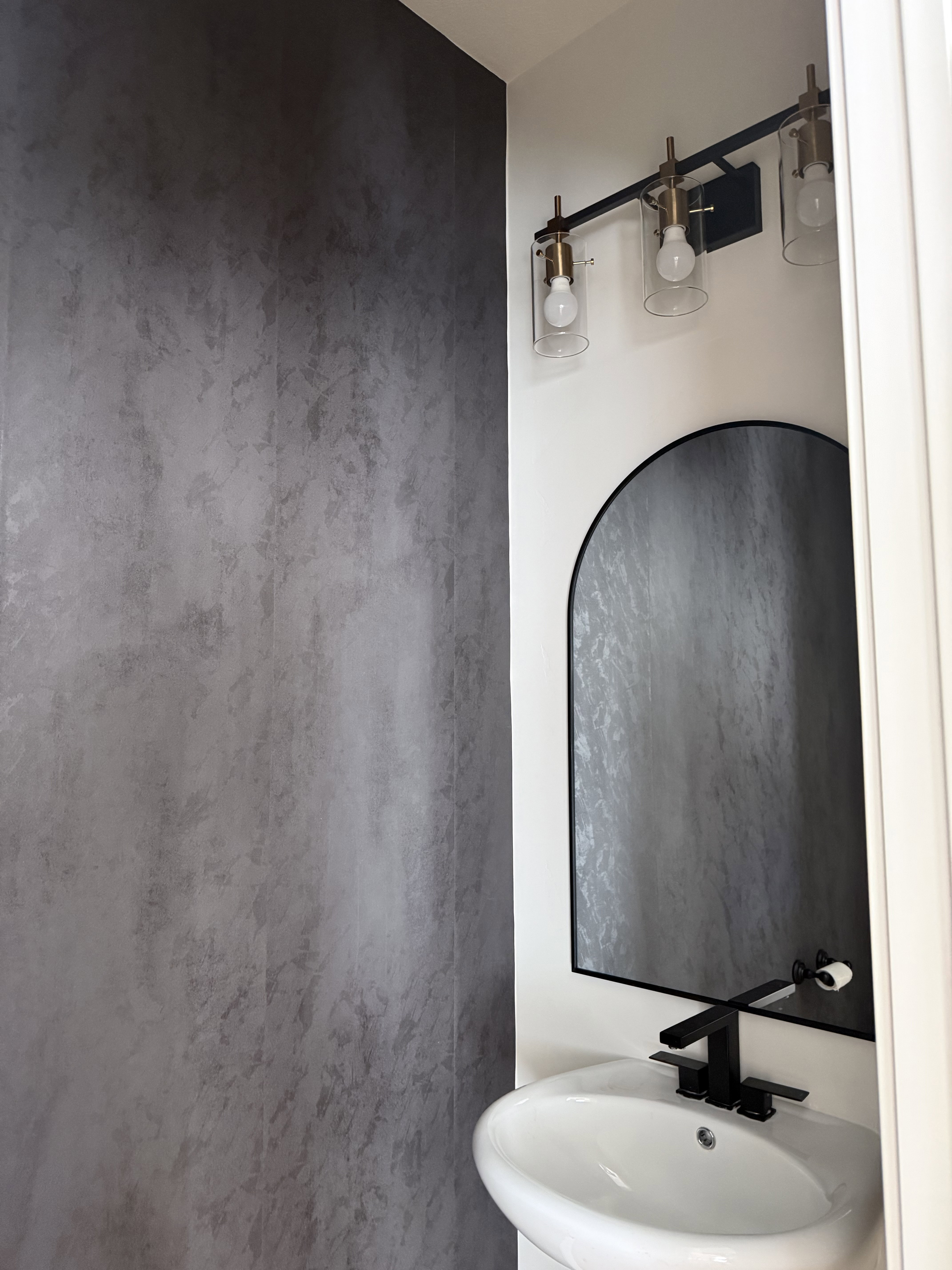 Modern powder room featuring a textured accent wall, black arched mirror, and a contemporary vanity light fixture with clear glass shades