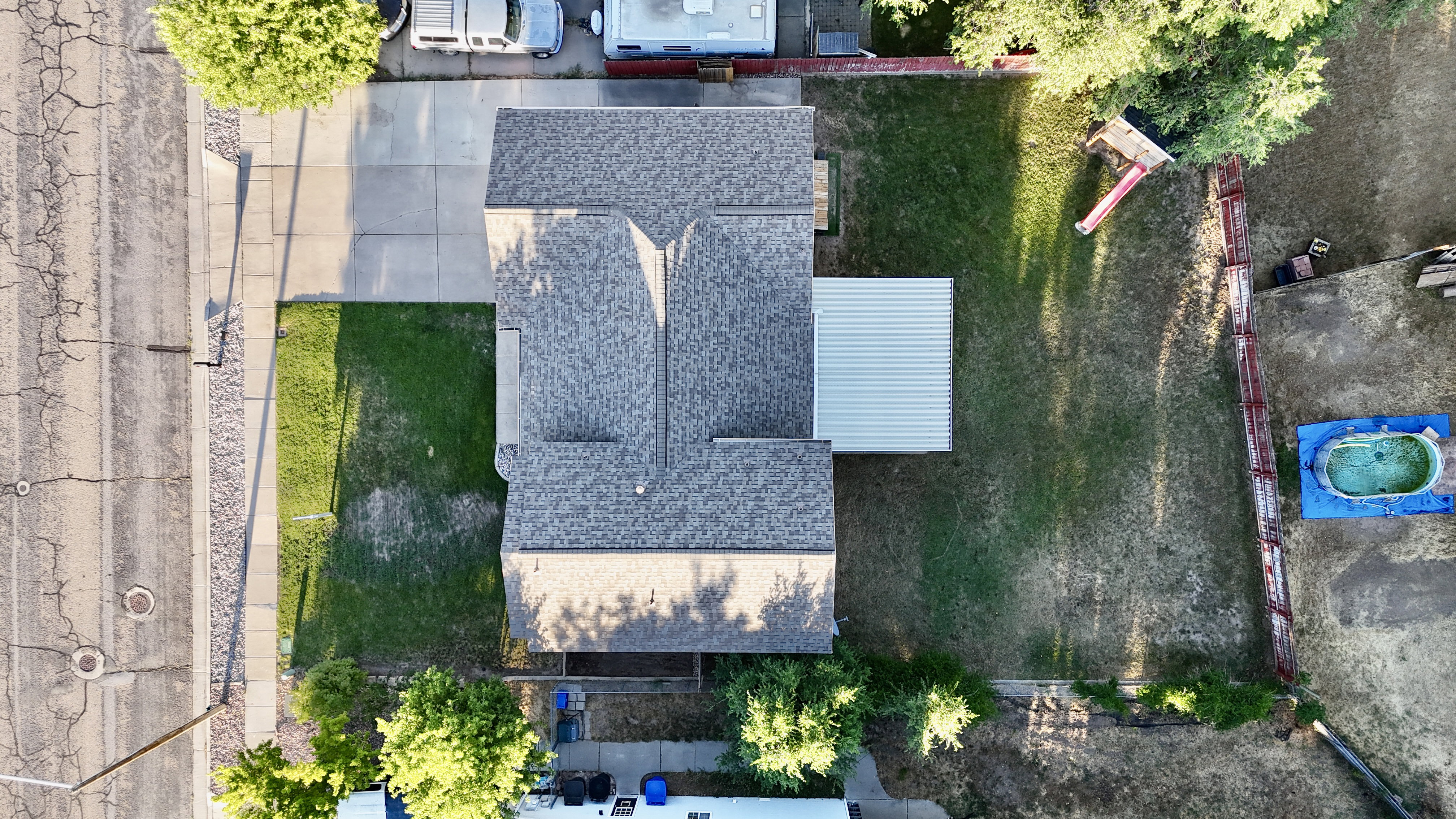 Aerial view showcasing a residential property featuring a gray shingle roof, a front lawn with a concrete driveway, a rear extension with a corrugated metal roof, and a fenced backyard