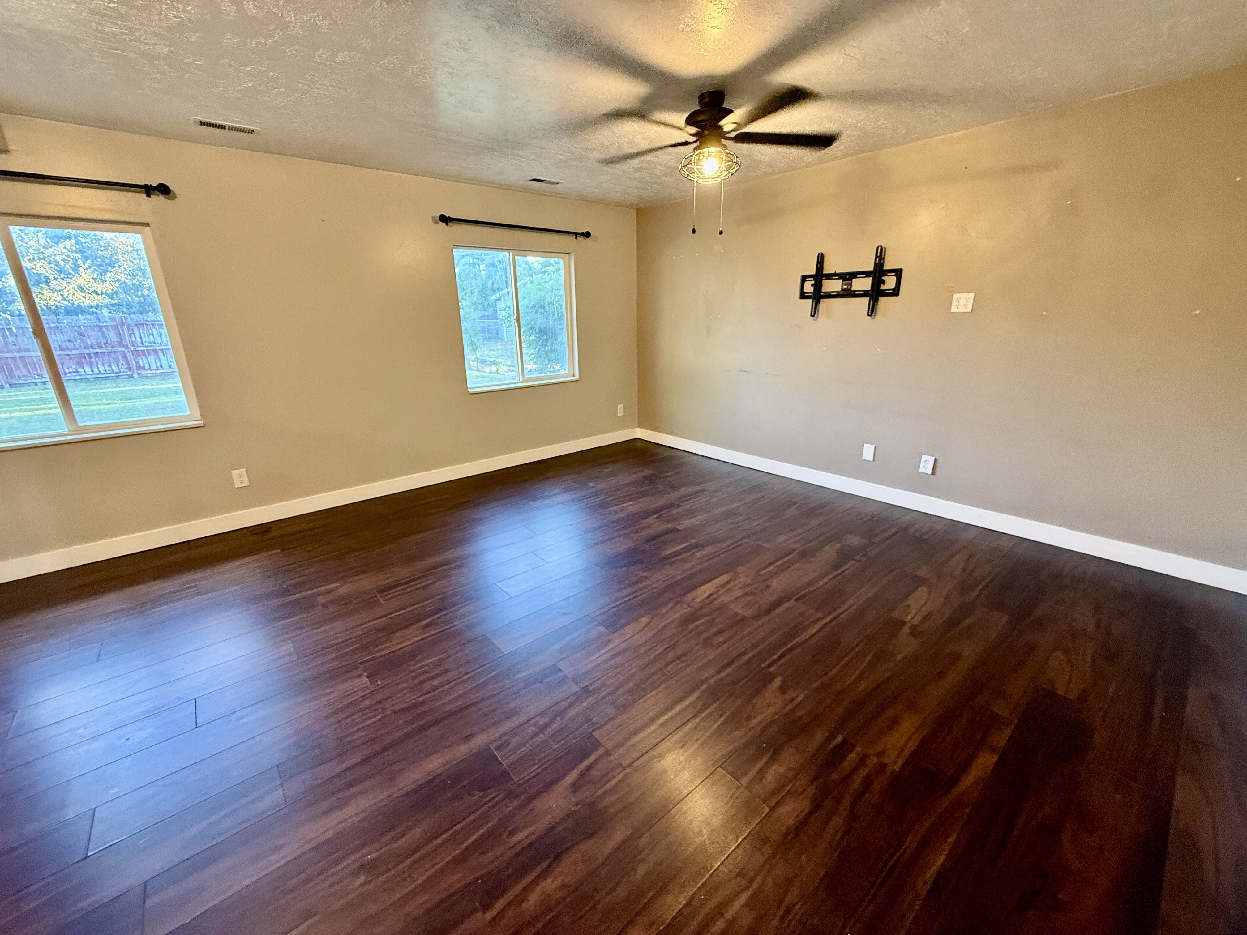 Spacious room featuring rich wood-finish flooring, neutral wall tones, and two windows providing outdoor visibility