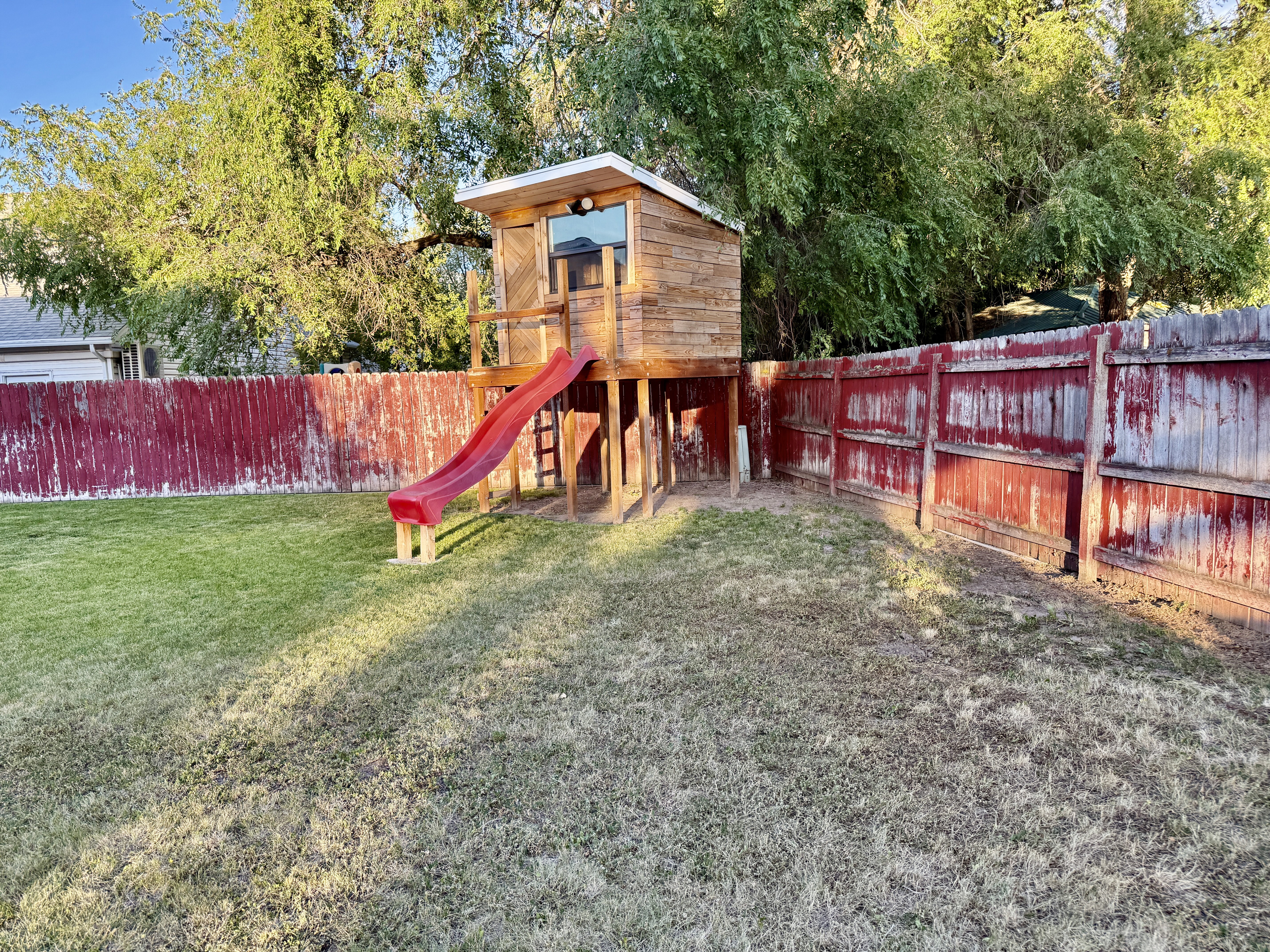 Fenced backyard featuring a wooden playhouse with a red slide, ladder access, and a rectangular window