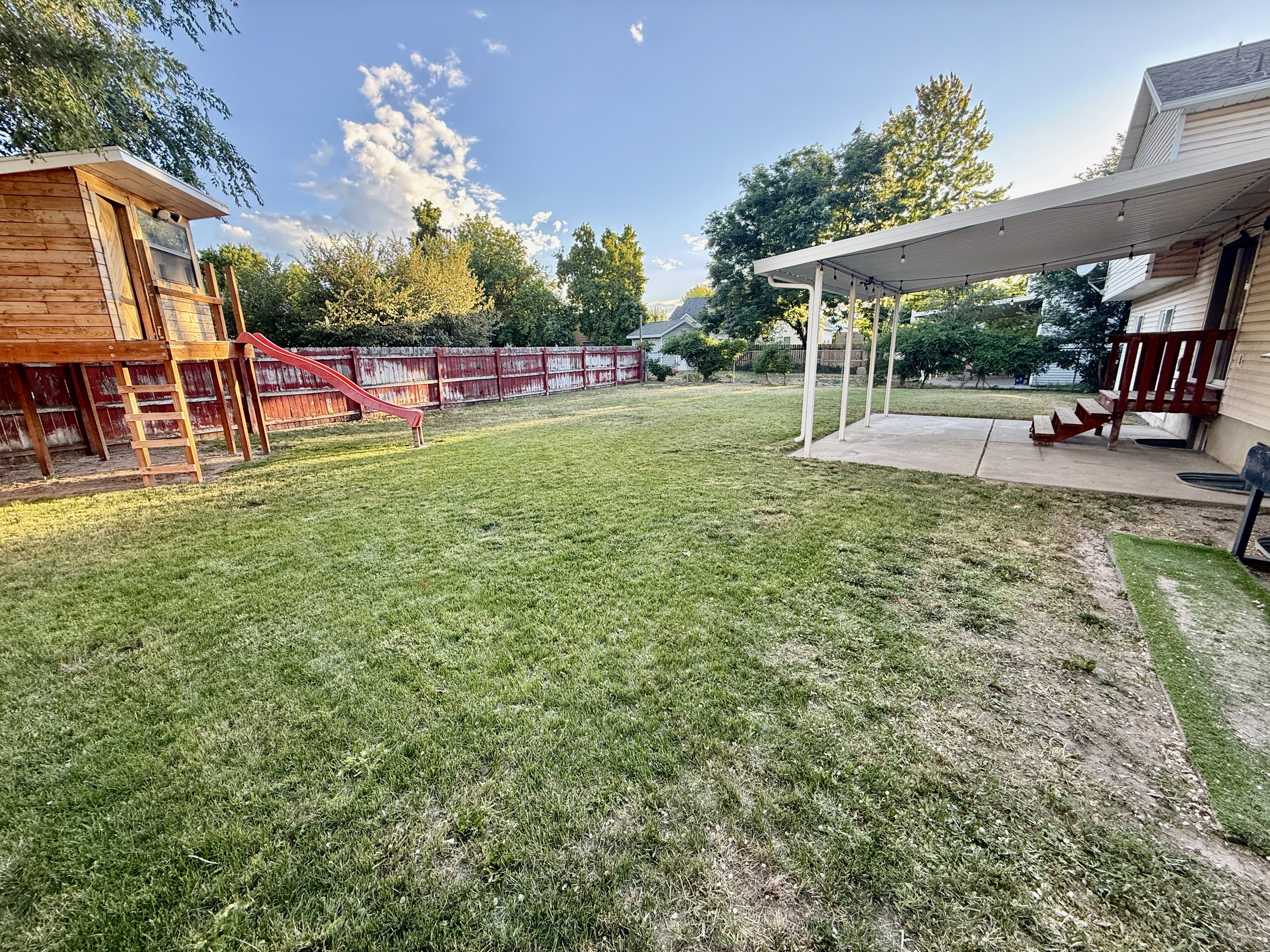 Expansive green yard featuring a covered patio, a red perimeter fence, and mature trees outlining the property