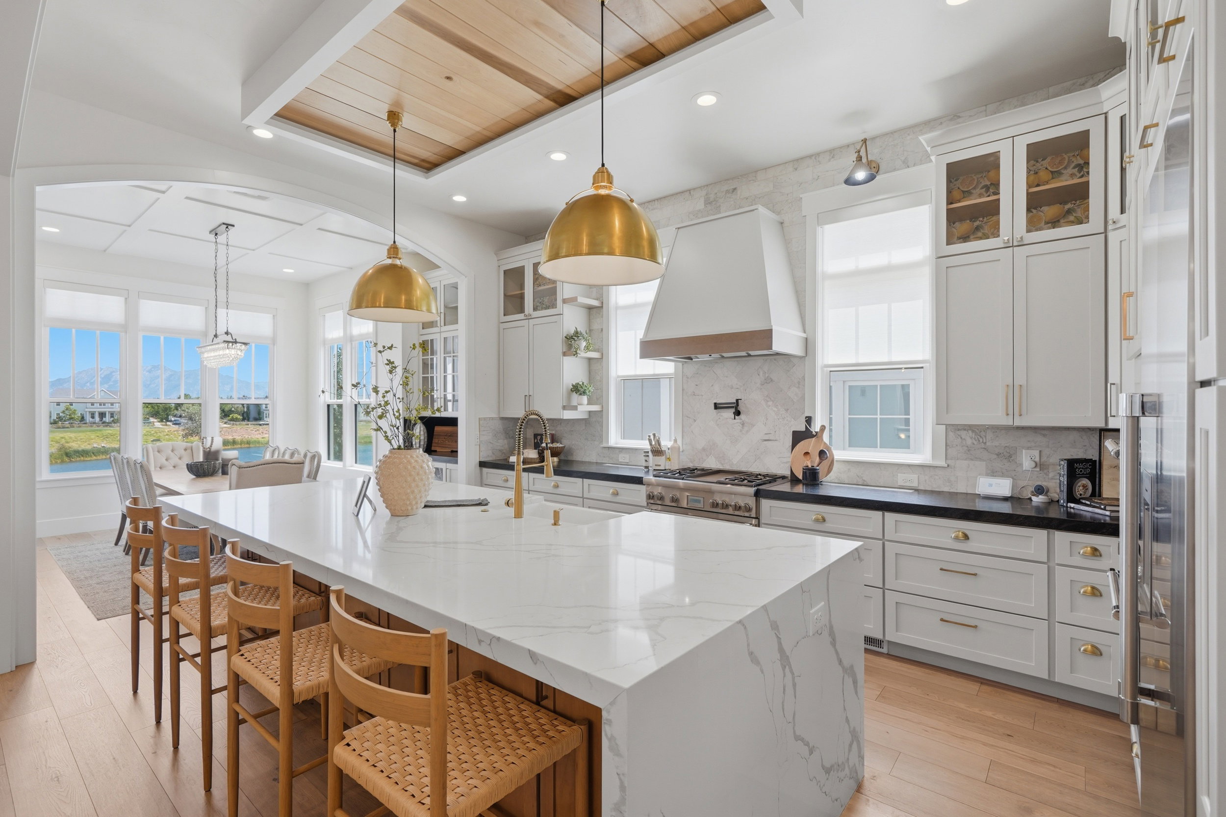 Gourmet kitchen featuring a large waterfall-edge island with seating, a coffered ceiling with wood-finish inlay, brass pendant lighting, a built-in range with a custom hood, and light-toned wood flooring