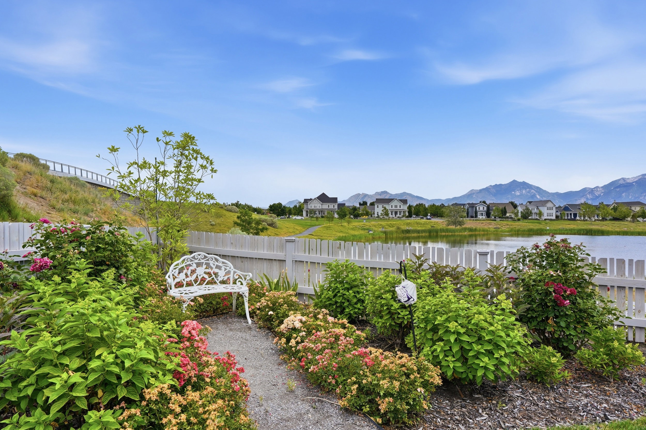 Lakeside garden area featuring a white picket fence, a gravel pathway, and a variety of blooming shrubs