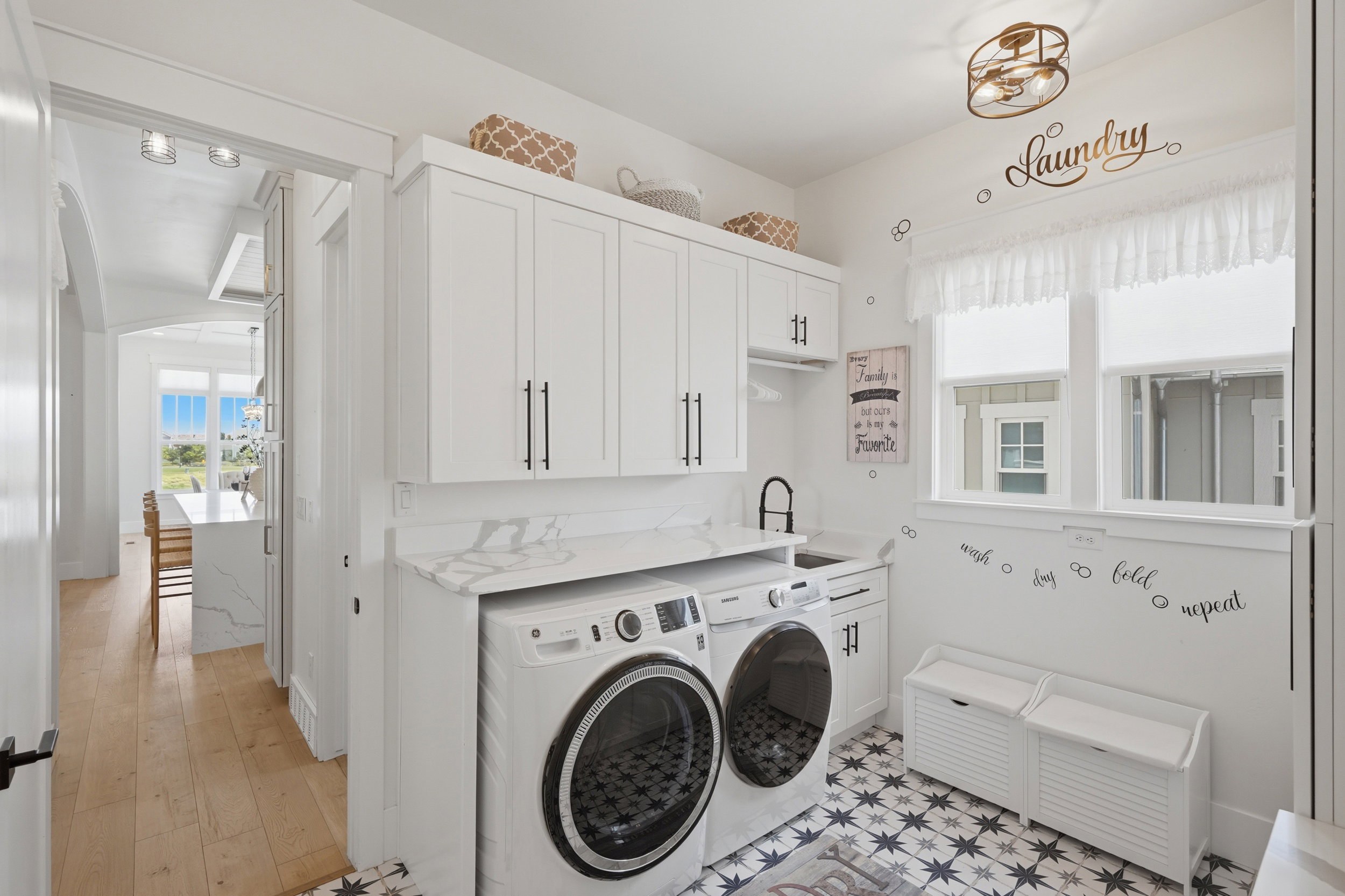 Laundry area featuring patterned tile flooring, white shaker cabinetry with dark hardware, a built-in sink with a matte black faucet, and a quartz-finish countertop