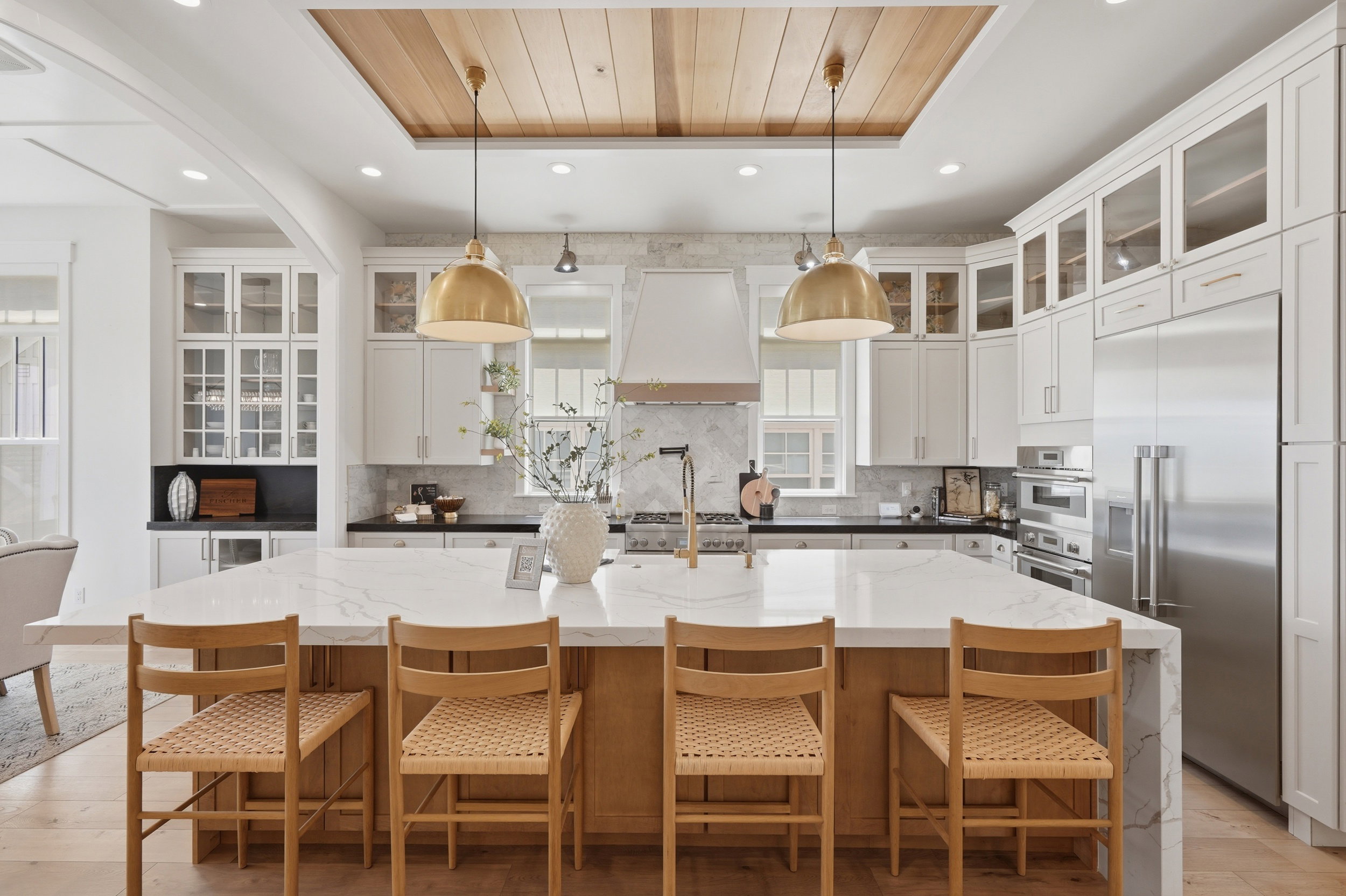 Gourmet kitchen featuring a large central island with seating, integrated stainless steel appliances, custom cabinetry with glass-front uppers, a patterned marble backsplash, and a wood-paneled ceiling accent
