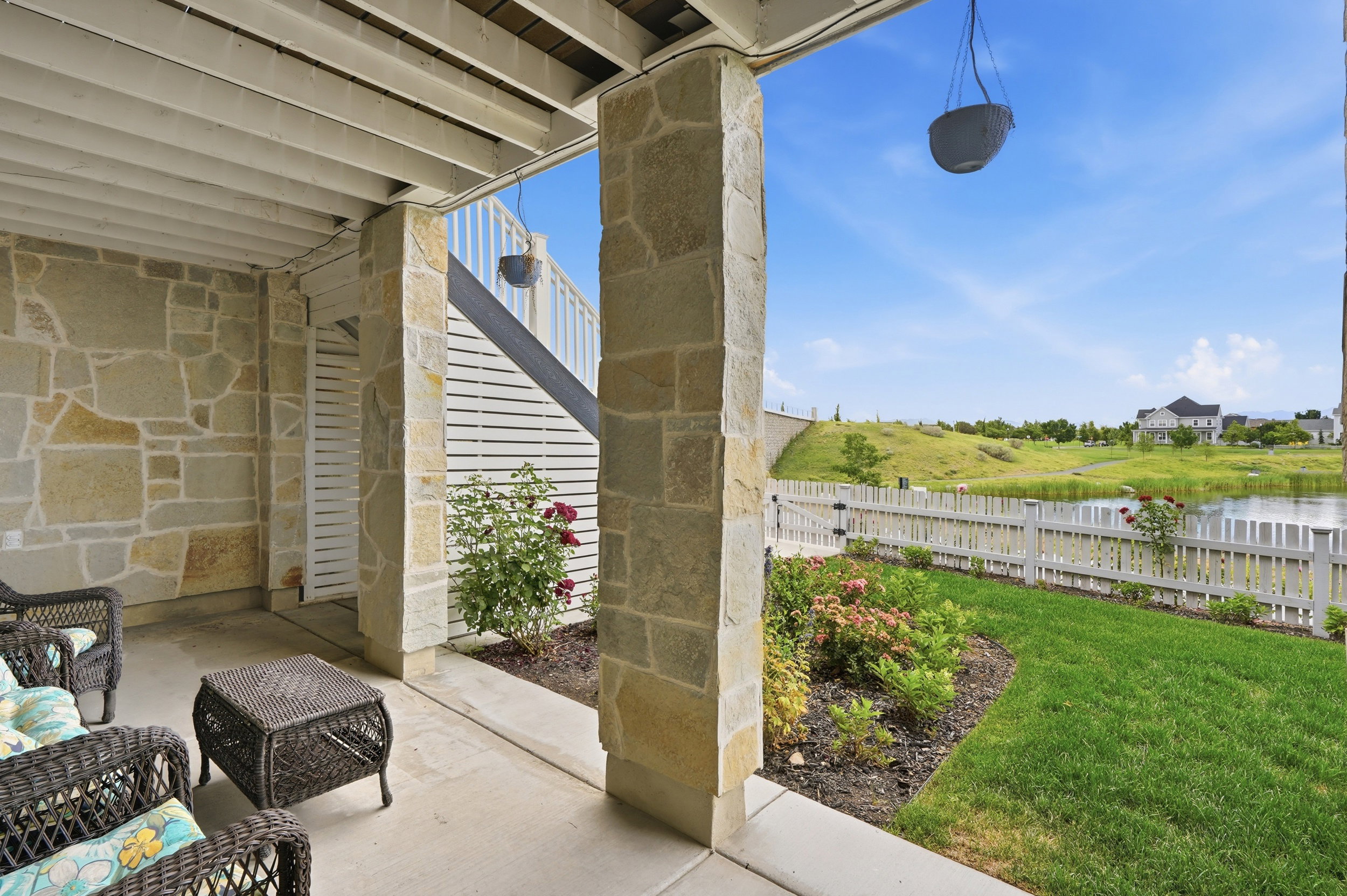 Covered patio with stone columns and wall detailing, featuring a concrete floor, a manicured lawn, and a white picket fence bordering a pond