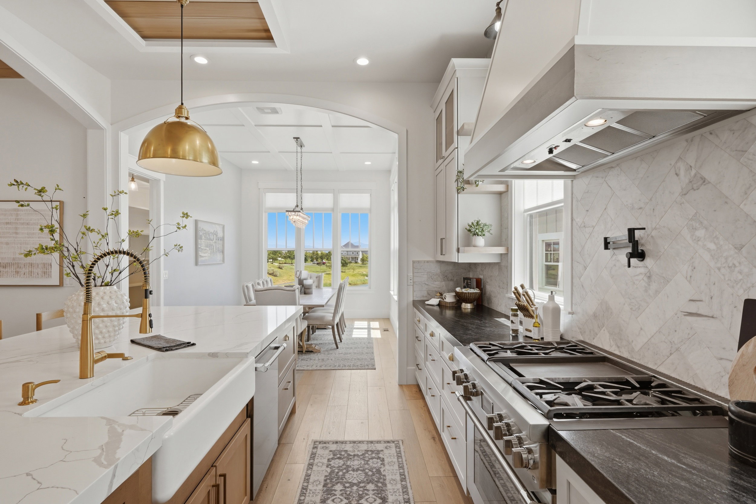 Gourmet kitchen featuring a white farmhouse sink, gold-tone fixtures, light wood-finish flooring, dark countertops, and a herringbone tile backsplash