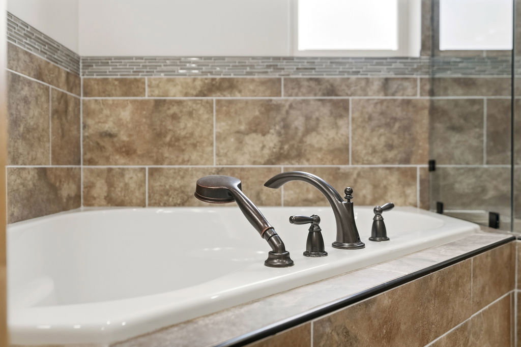 Soaking tub featuring a deck-mounted bronze faucet with a handheld sprayer, surrounded by large format tile with a decorative mosaic accent band
