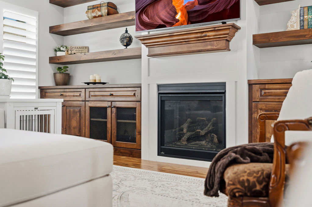 Central fireplace with a wood mantel, flanked by built-in wood cabinetry, floating wood shelves, and white plantation shutters