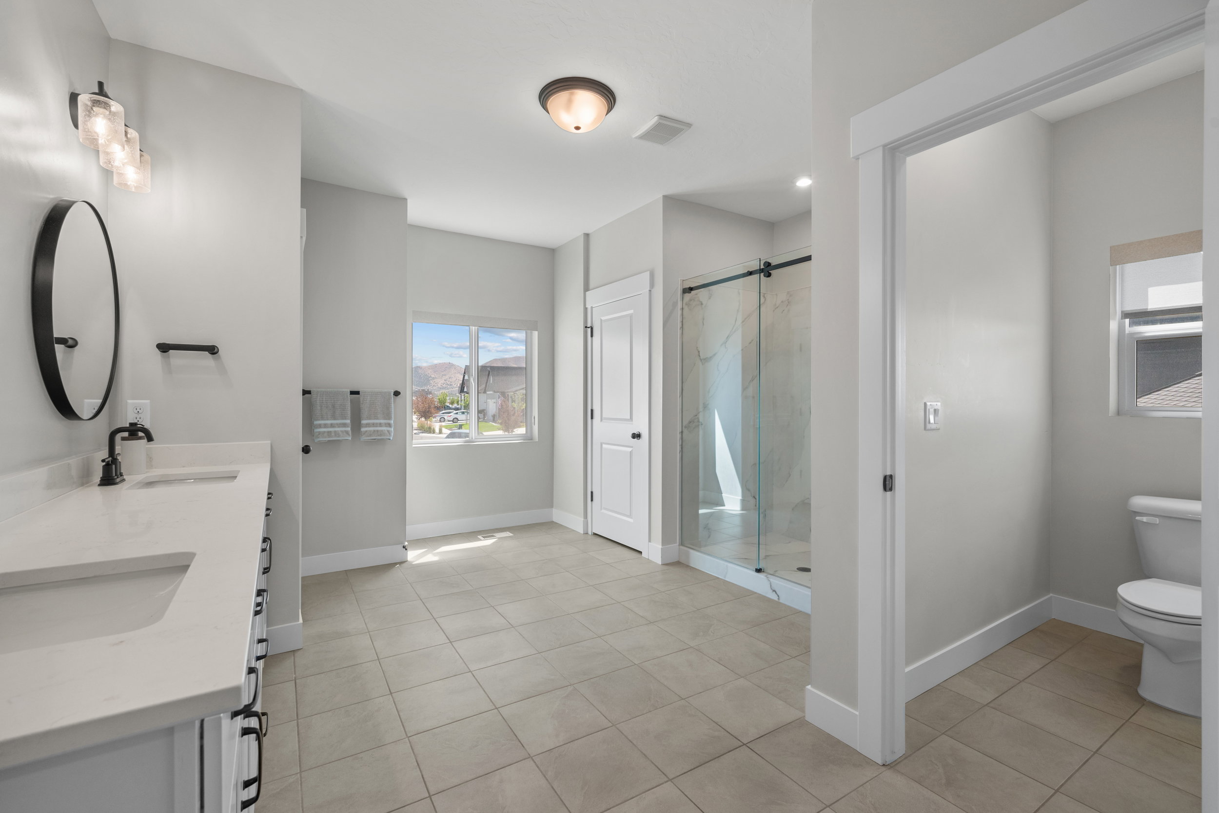 Spacious bathroom featuring a double vanity with stone-finish countertops, a glass-enclosed shower, neutral tile flooring, and a separate water closet