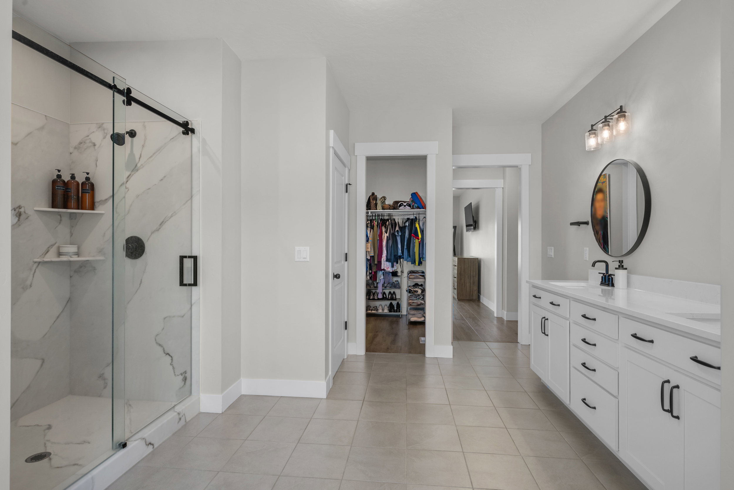 Spacious bathroom featuring a frameless glass shower with marble-look tile, a double vanity with white cabinetry and dark hardware, and large format floor tile