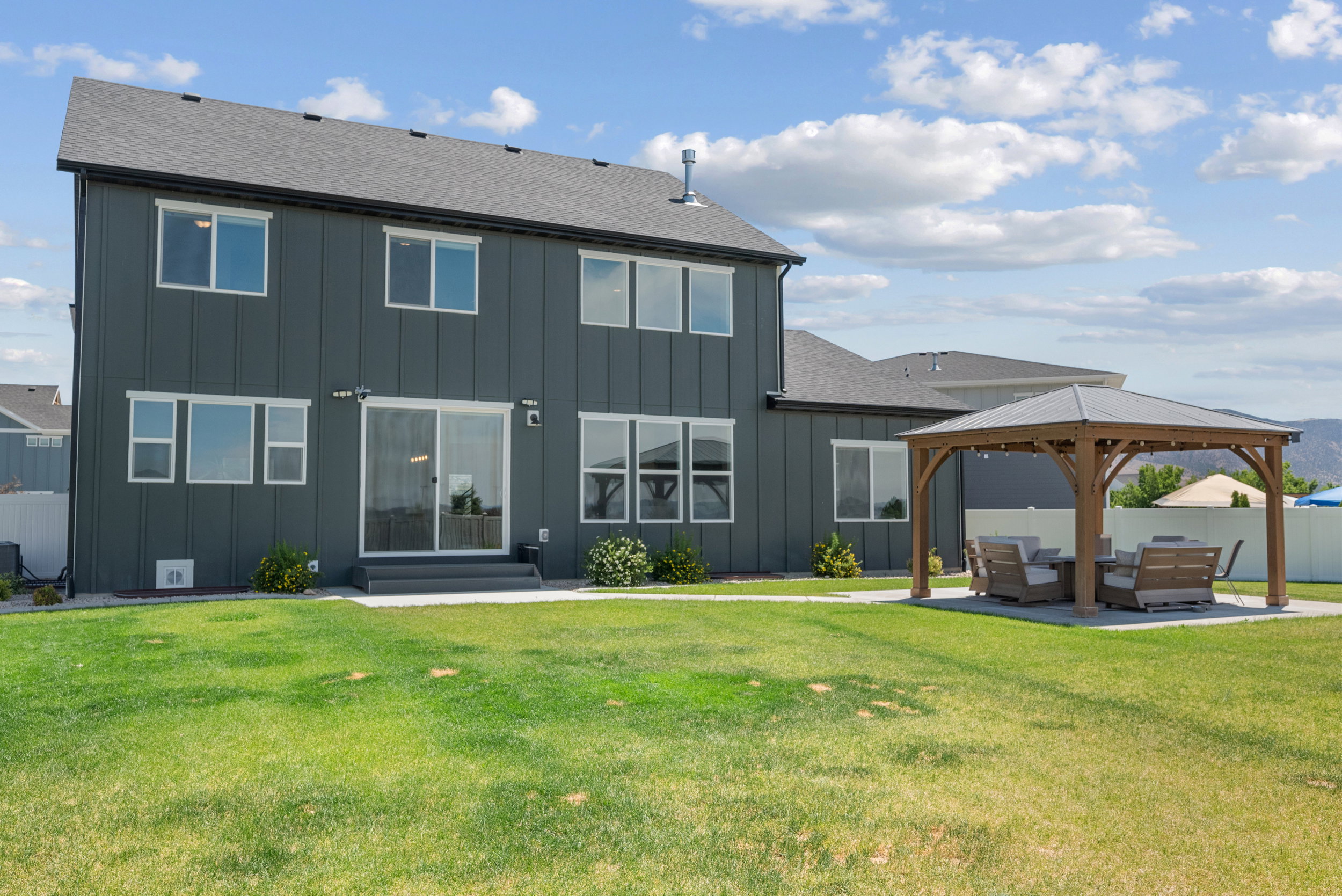 Expansive backyard featuring a modern gray siding exterior, a dark gray asphalt shingle roof, white-framed windows, a sliding glass patio door, and a built-in wooden gazebo with a gray roof situated on a concrete pad