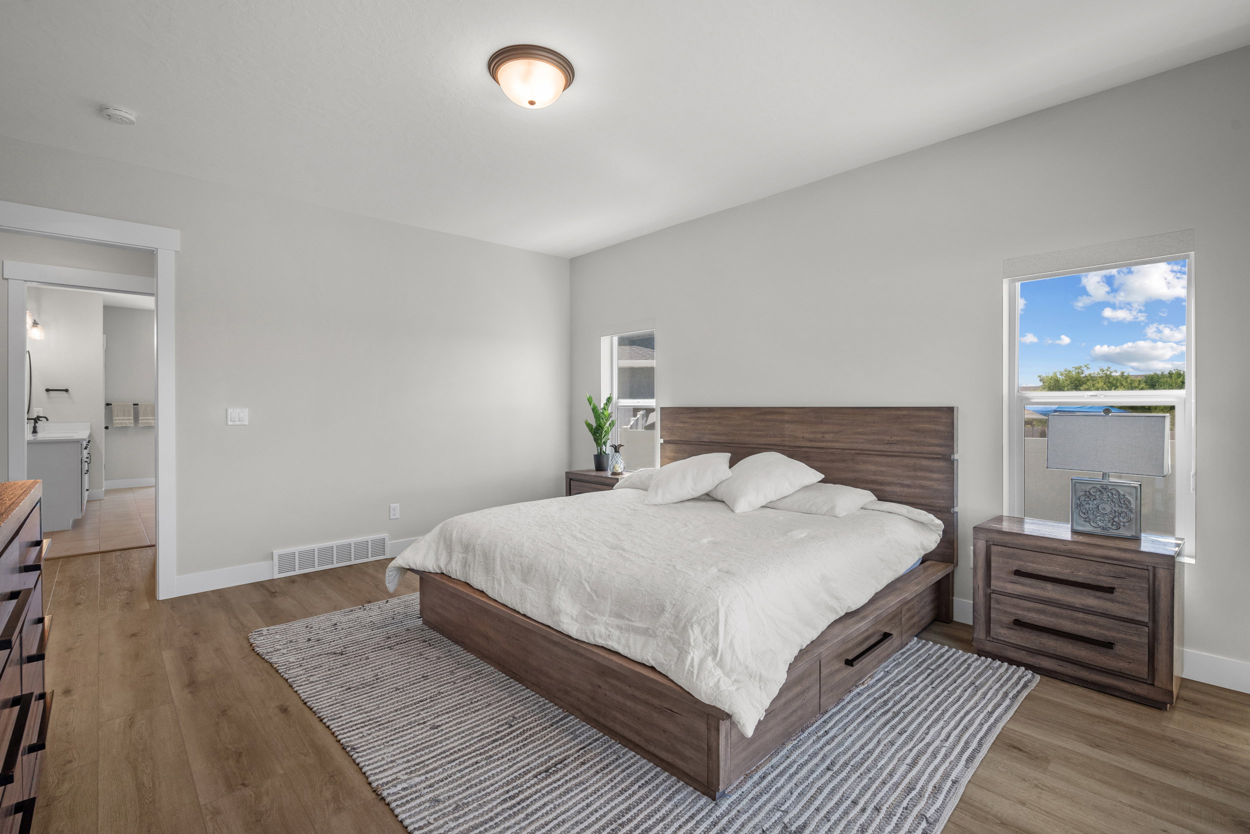 Spacious bedroom featuring light neutral wall tones, wood-finish flooring, a contemporary ceiling light fixture, and a large window providing natural light