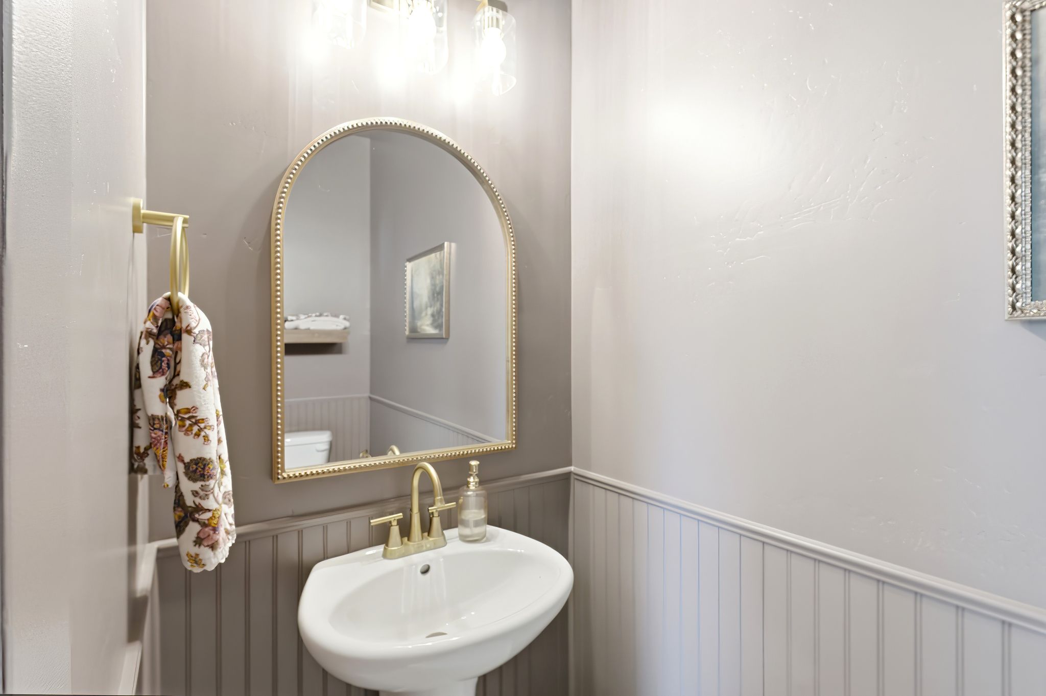 Pedestal sink with brass-finish faucet, framed by wainscoting and a decorative arched mirror