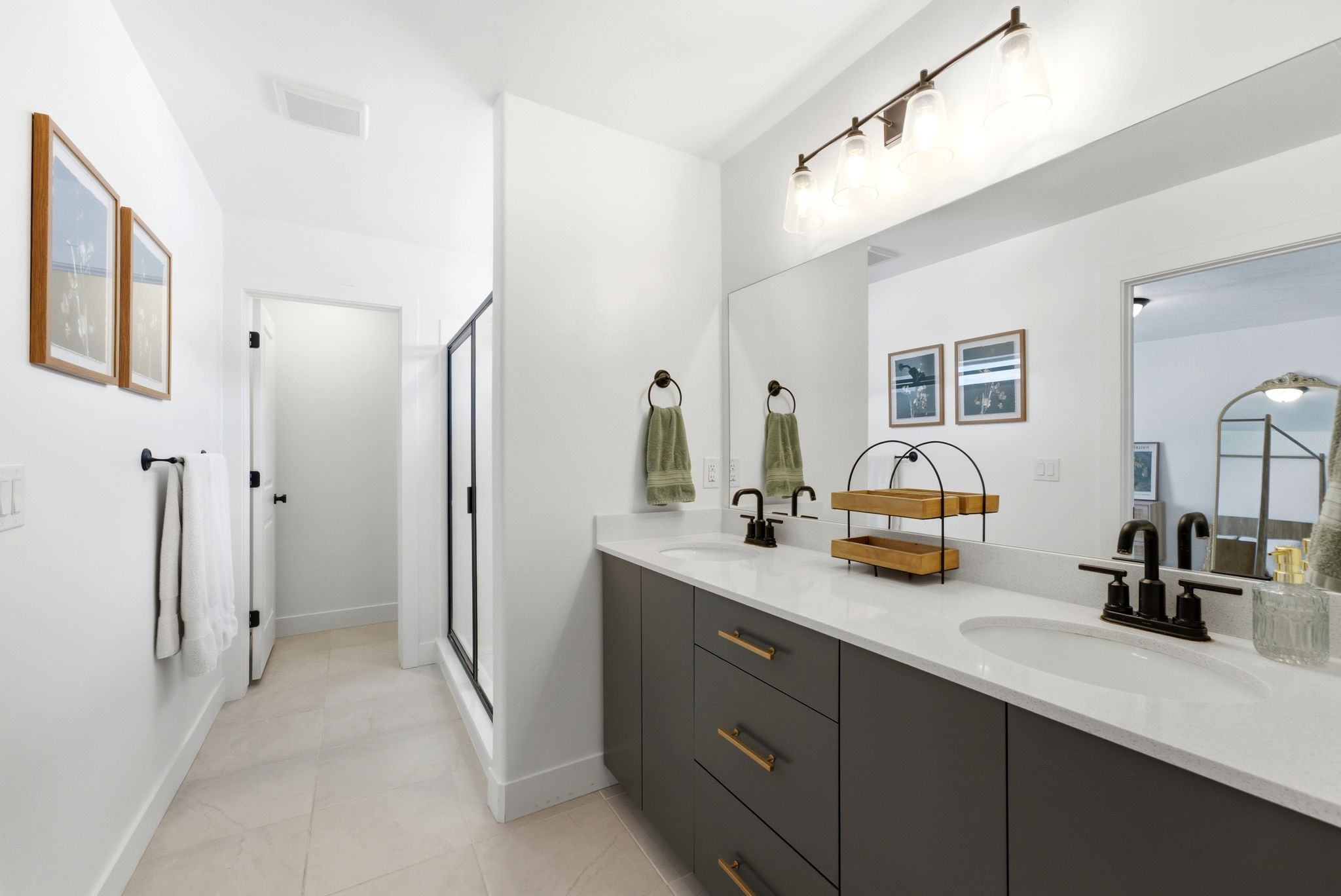 Dual vanity with white countertops and matte black fixtures, dark gray cabinetry with gold-tone hardware, full-width mirror, and tiled flooring