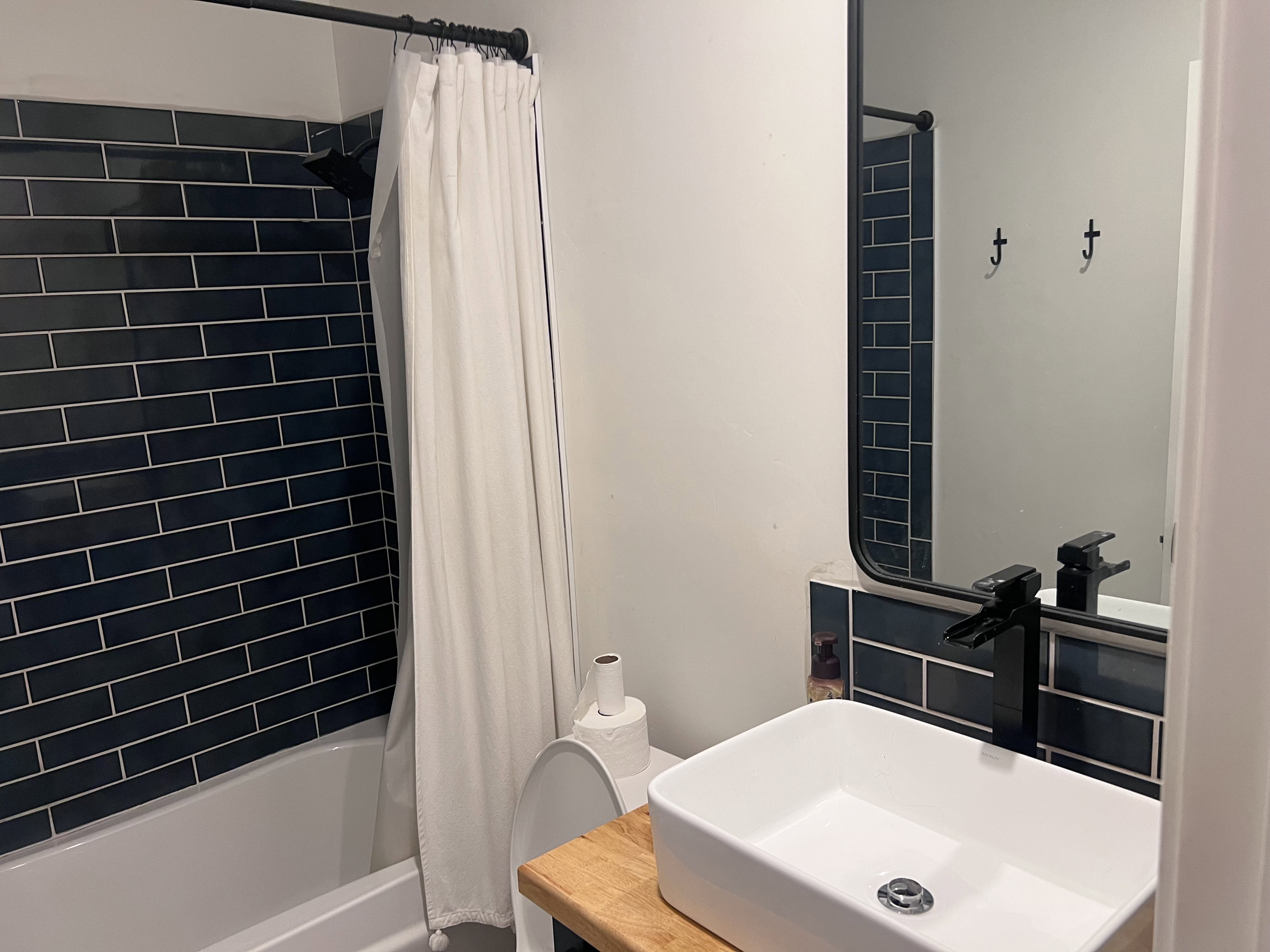 Bathroom featuring a vessel sink with a matte black faucet, a rectangular mirror, and a built-in tub with dark blue subway tile surround