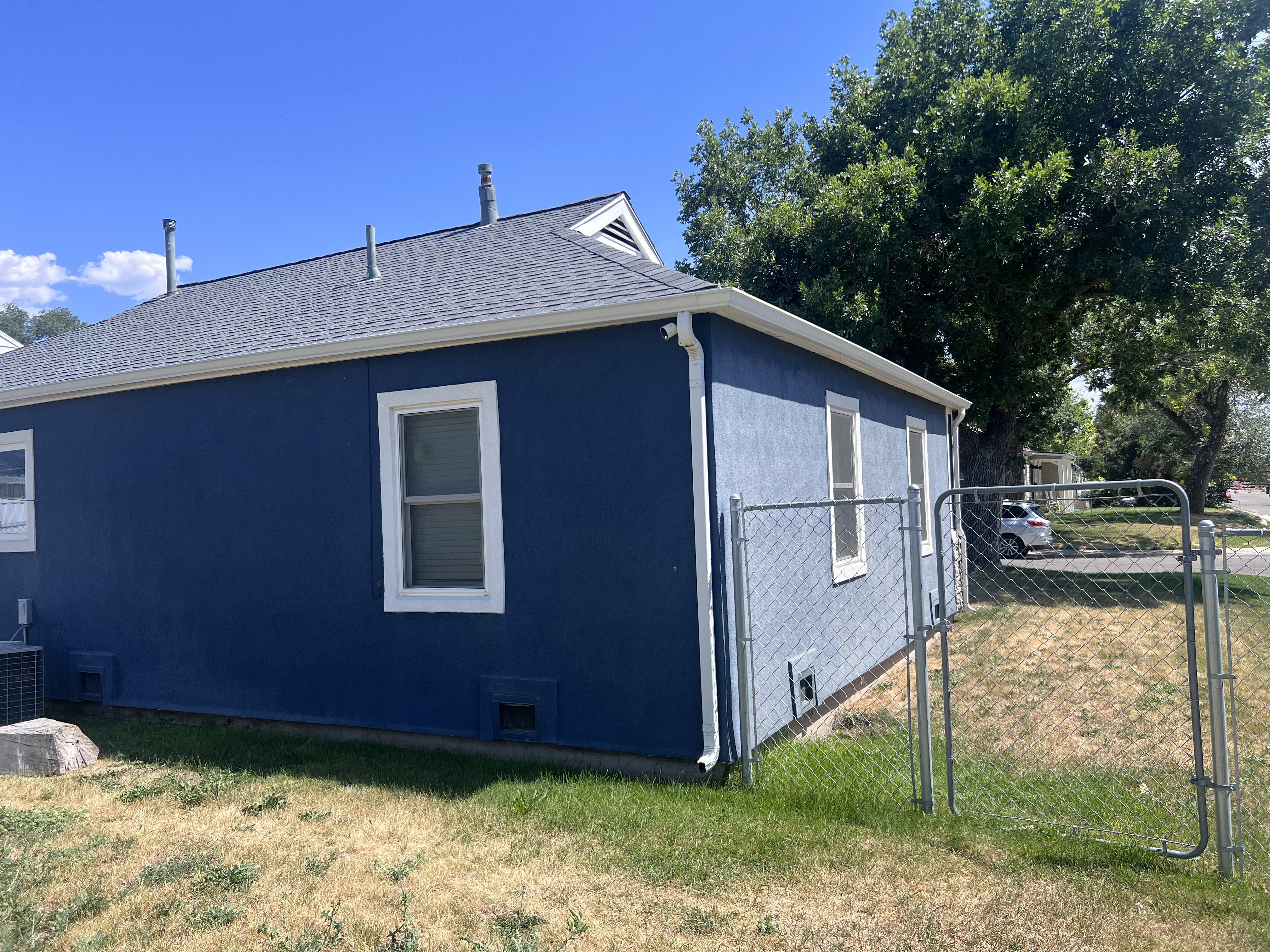 Exterior featuring dark blue siding, white trim windows, gray shingle roofing, white gutters, and a chain-link fence